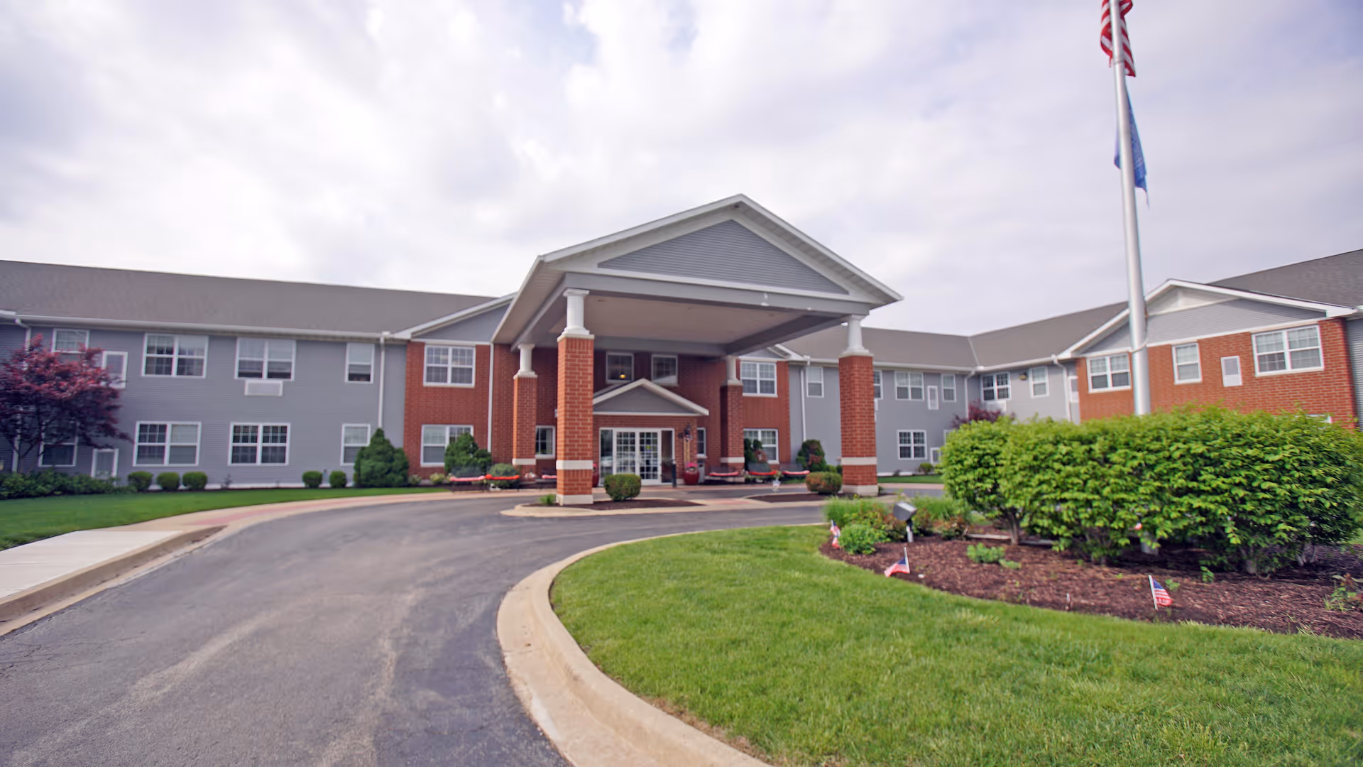 Entrance of a two-story brick and siding senior living building with a covered porte-cochère and landscaped driveway.