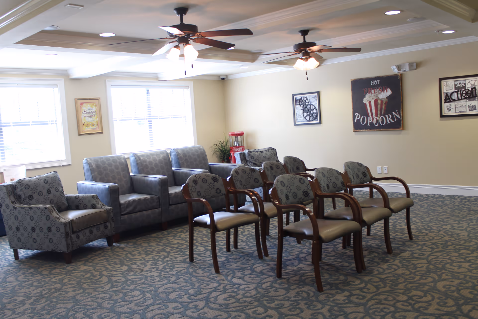 A well-lit common room with multiple chairs arranged in rows facing a blank wall. The room has patterned carpet, two ceiling fans with lights, and several framed pictures on the walls, including one with a popcorn theme. There are windows letting in natural light and a popcorn machine in the corner.