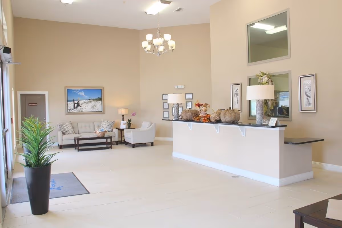 A bright and spacious reception area with a beige color scheme. There is a long reception desk decorated with lamps and autumn-themed decor. To the left, there is a seating area with a sofa, armchair, coffee table, and a framed beach photograph on the wall. A tall potted plant is near the entrance door, and the floor is tiled in a light color.