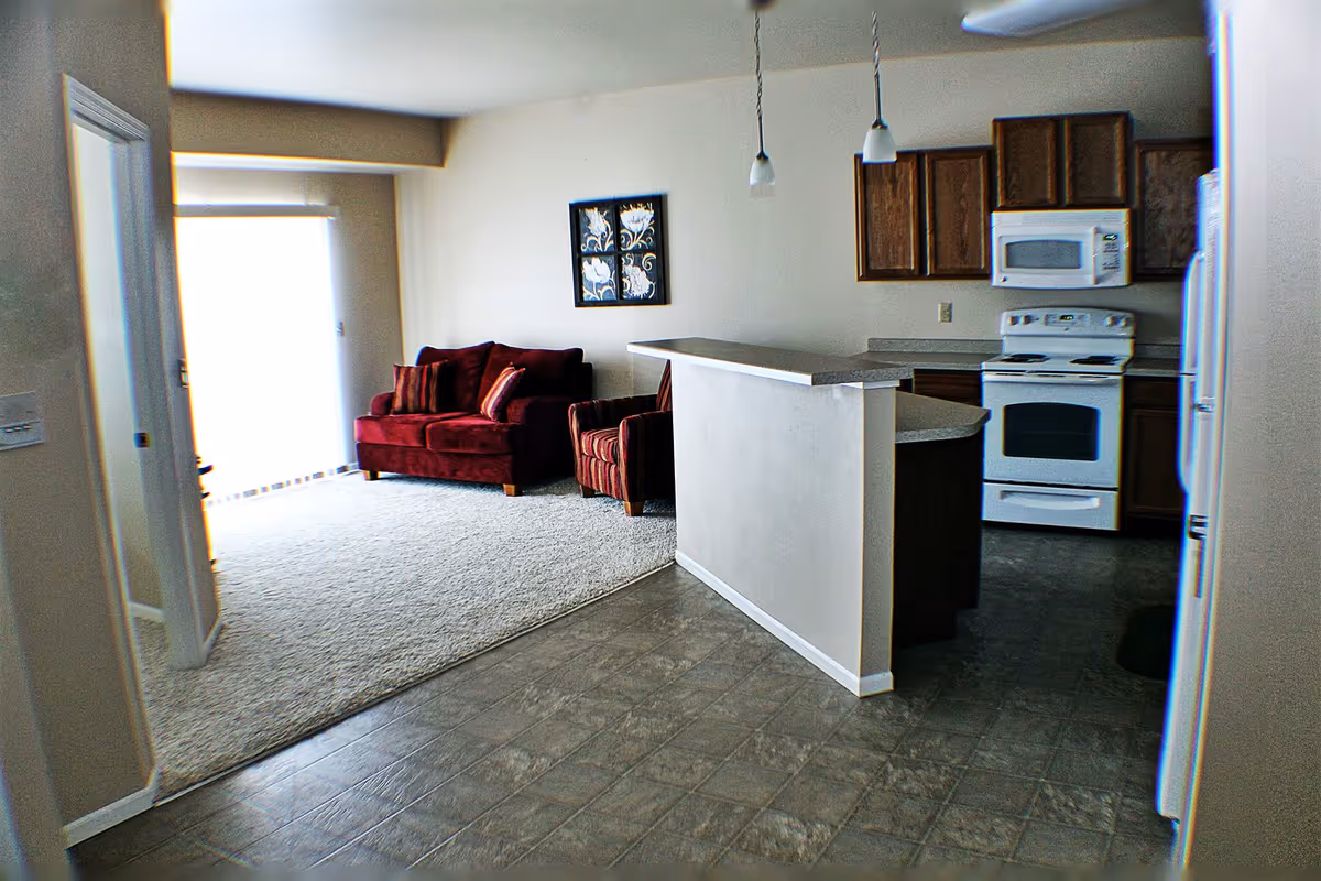 Open-plan living area showing a red loveseat and chair on carpet next to a kitchen with a breakfast bar, wooden cabinets, and white appliances.