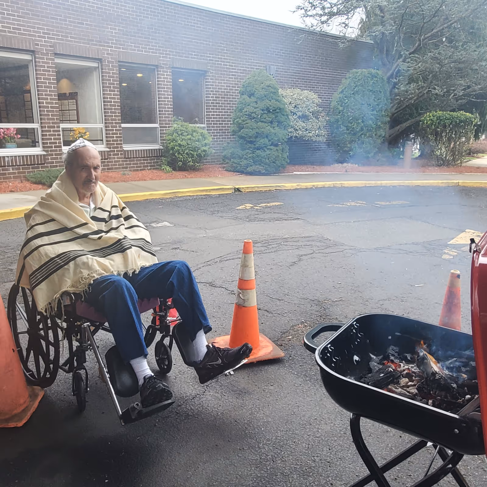 An elderly man in a wheelchair wrapped in a tallit (Jewish prayer shawl) and wearing a kippah, sitting outside near a small fire pit with burning coals. The setting is a paved area outside a brick building with some bushes and windows in the background. There are orange traffic cones placed around the area.