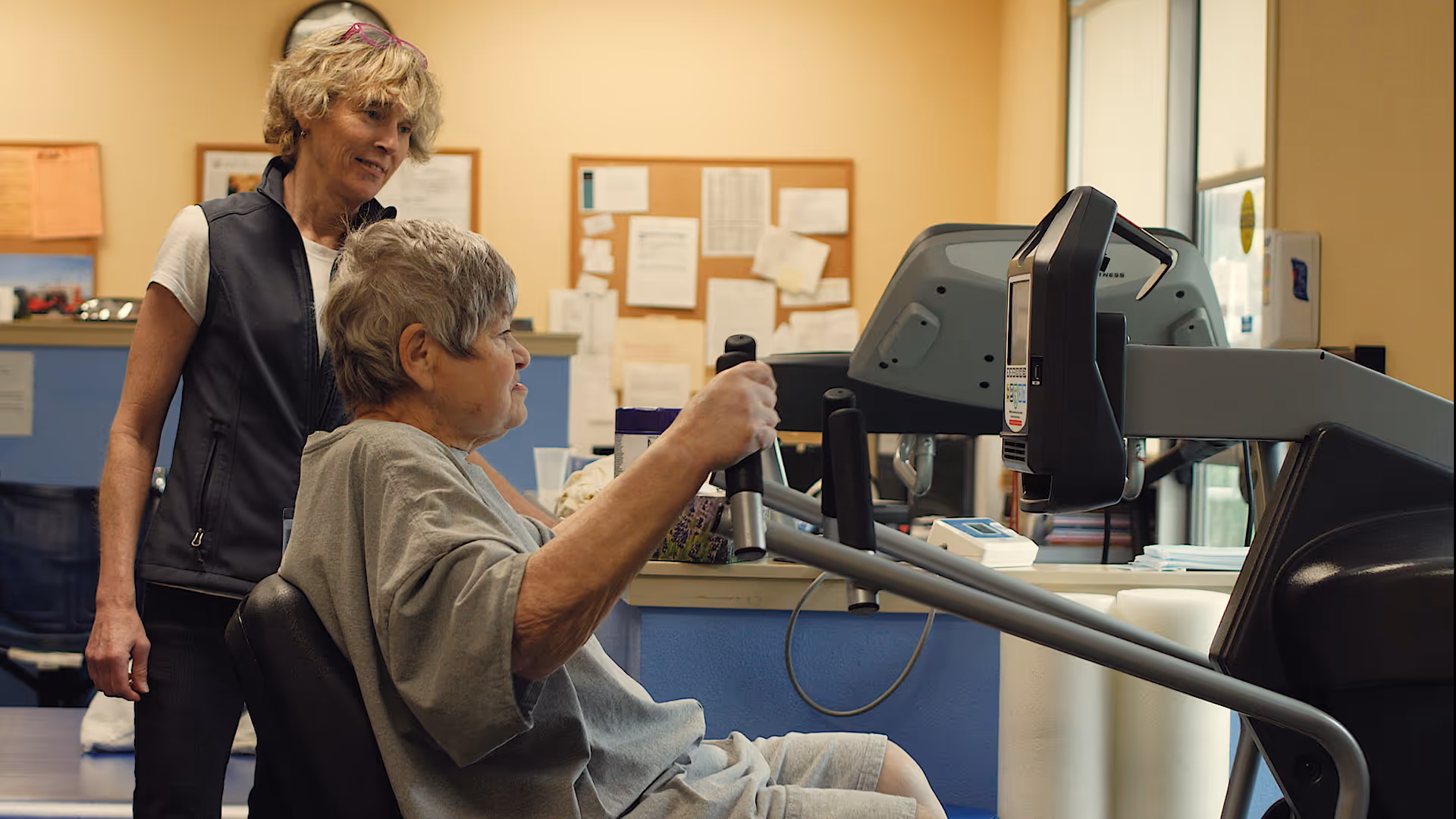 An elderly woman using a seated exercise machine in a fitness or rehabilitation room, assisted by a standing caregiver or trainer. The room has a bulletin board with papers pinned on the wall in the background.