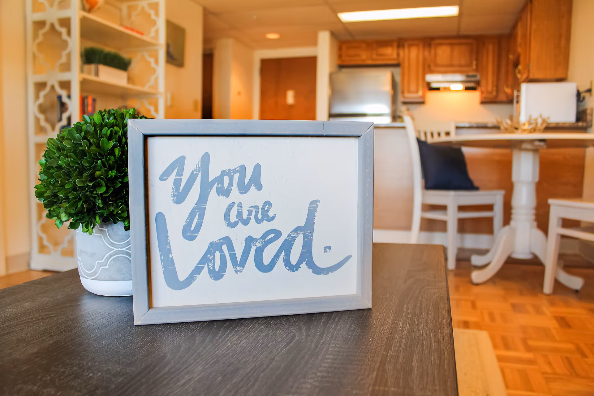 A framed sign on a dark wooden table with the words 'You are loved' written in blue cursive. Next to the sign is a small potted green plant. In the background, there is a kitchen and dining area with wooden cabinets, a refrigerator, a dining table, and chairs.