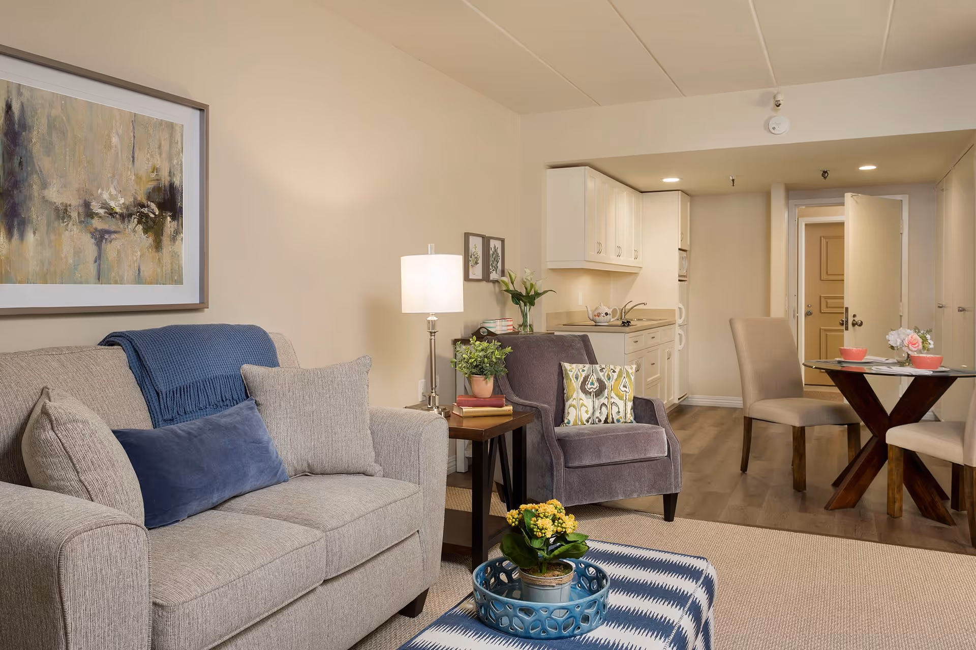 A cozy living room area with a beige sofa adorned with blue and gray pillows, a gray armchair with a patterned cushion, a side table with a lamp and potted plant, and a blue and white patterned ottoman with a small potted plant on a tray. In the background, there is a small dining area with a round glass table and two beige chairs, and a kitchenette with white cabinets and a countertop. The walls are light-colored and decorated with framed artwork.