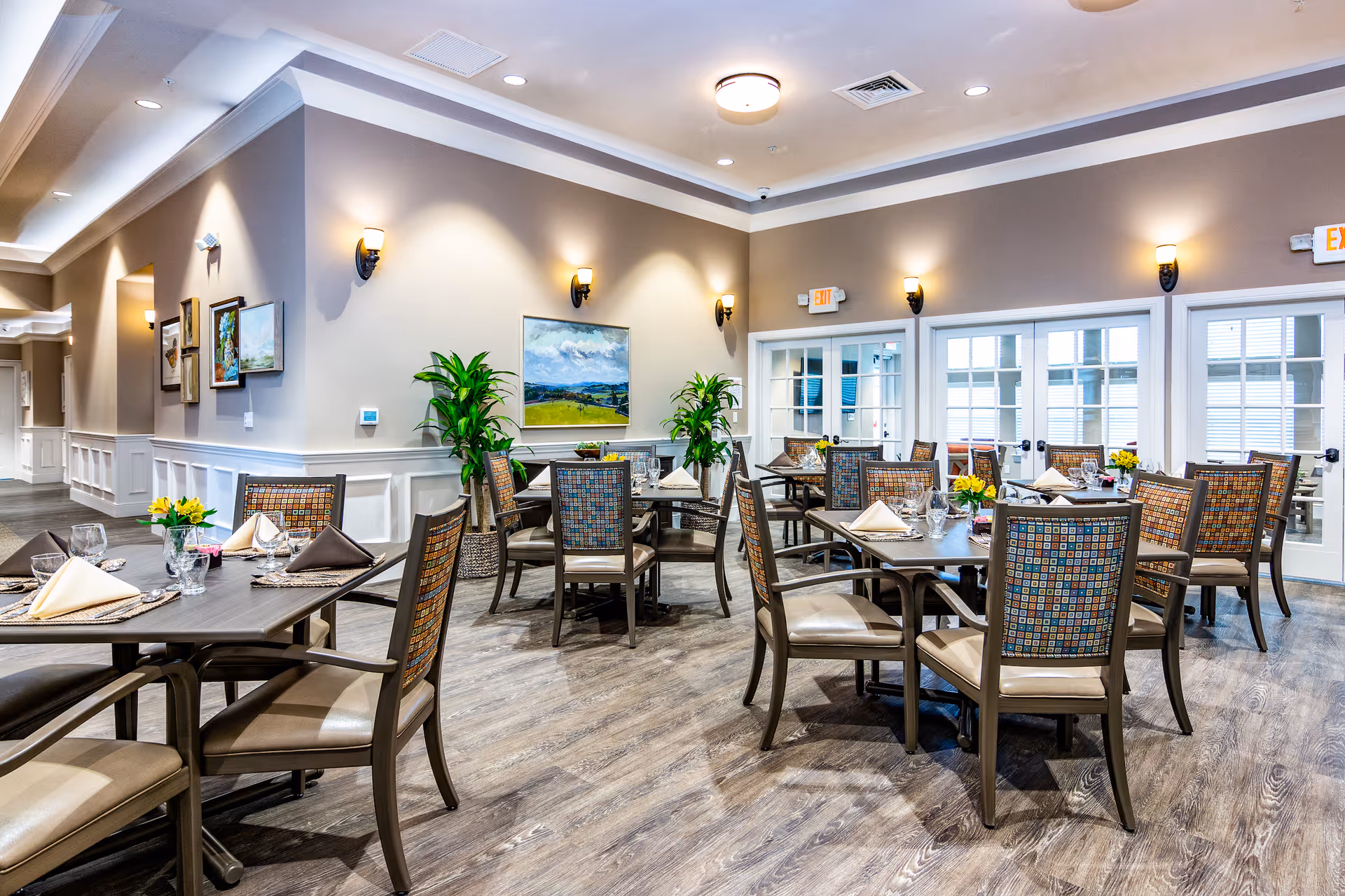 A bright and spacious dining room in an assisted living facility with multiple tables set with napkins, glasses, and small flower arrangements. The room features wooden flooring, beige walls with white wainscoting, wall sconces, framed artwork, and large windows with glass doors allowing natural light to enter. There are several chairs with patterned upholstery around each table.