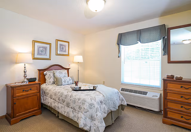 A cozy bedroom with a single bed featuring floral bedding and multiple pillows. There is a wooden nightstand with a lamp and clock radio on the left side of the bed, and a wooden dresser with a mirror on the right side of the room. A window with blinds and a valance lets in natural light, and a floor lamp stands in the corner near the bed.