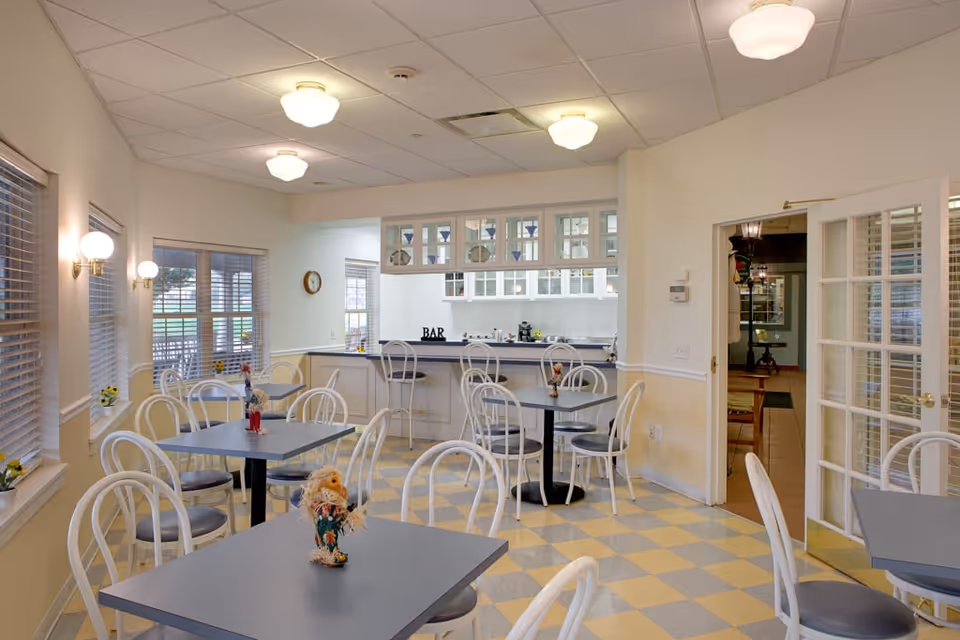 A bright dining area with several square tables and white chairs with gray seats. The floor has a yellow and gray checkered pattern. There are windows with blinds on the left wall, and a bar area with stools and glass cabinets in the background. The room is well-lit with ceiling lights and wall sconces. A doorway with glass-paneled double doors leads to another room.