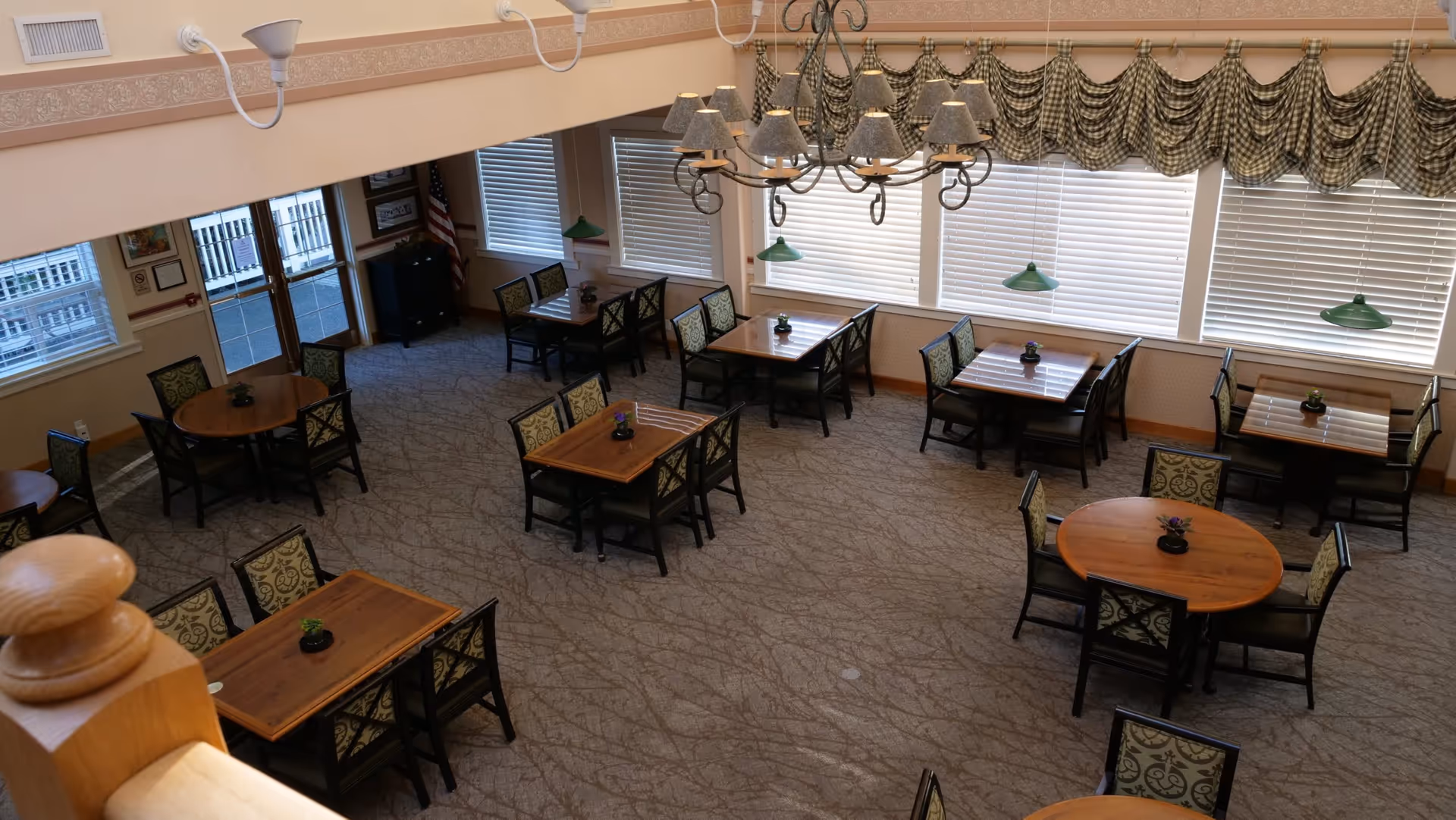 Interior view of a dining room with multiple wooden tables and chairs arranged neatly on a patterned carpet. The room features large windows with blinds and checkered valances, a chandelier with multiple lampshades, and green pendant lights hanging above some tables. There is an American flag in the corner near the entrance door.