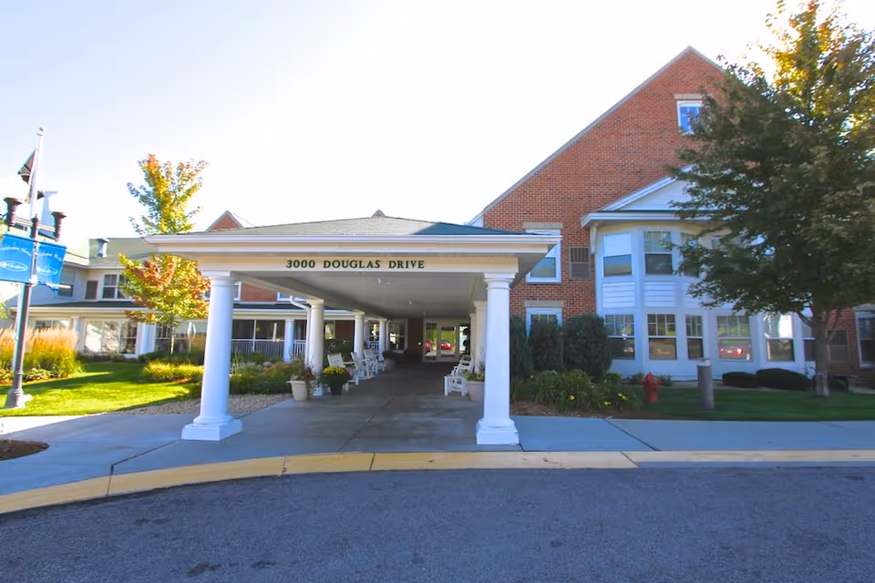 Front exterior view of a senior living facility with a covered entrance supported by white columns. The building is made of red brick with white trim and has several windows. There are trees and landscaping around the entrance, and a sign above the entrance reads '3000 DOUGLAS DRIVE'.