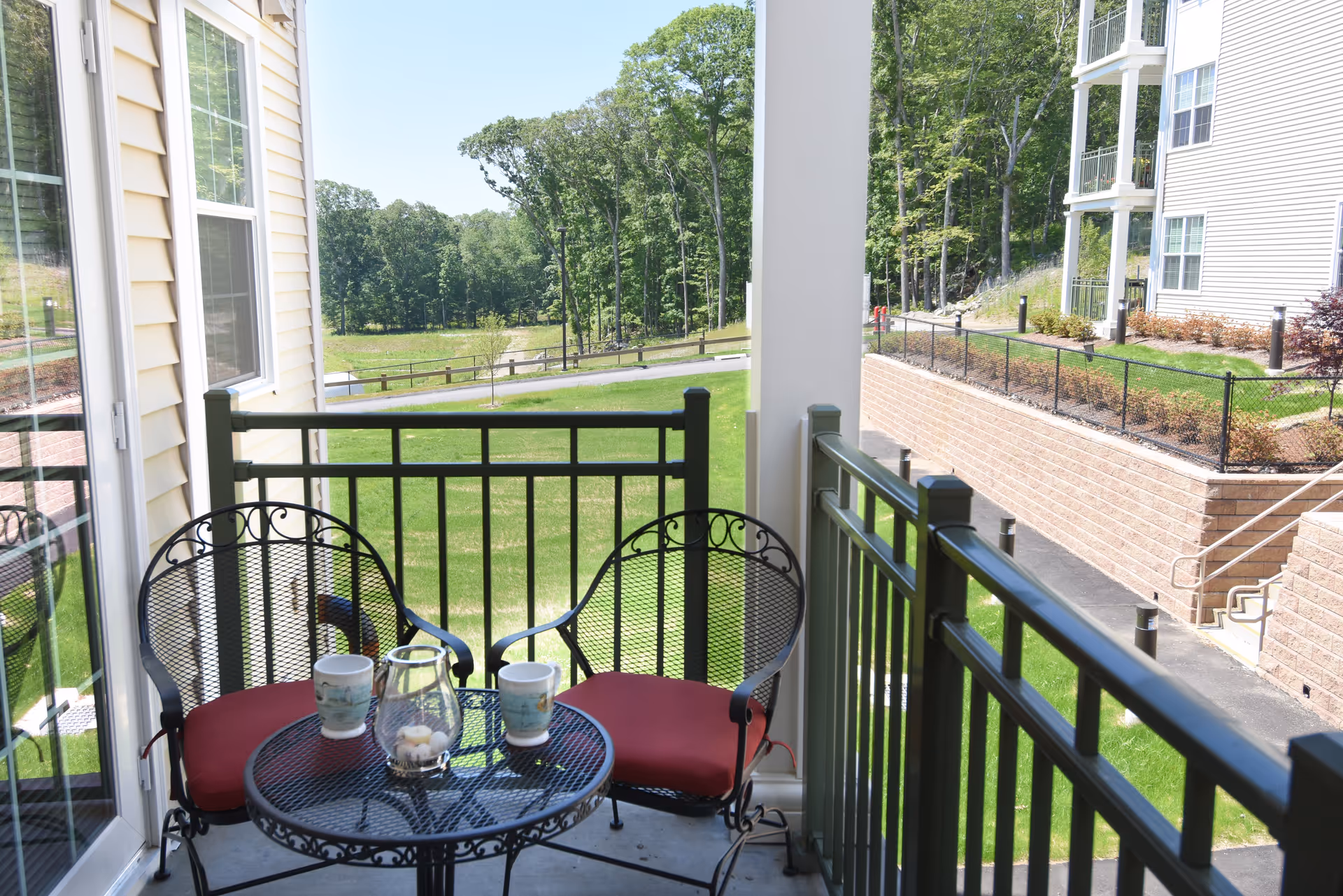 Small furnished balcony with two red-cushioned chairs and a round table overlooking a grassy lawn and neighboring building.