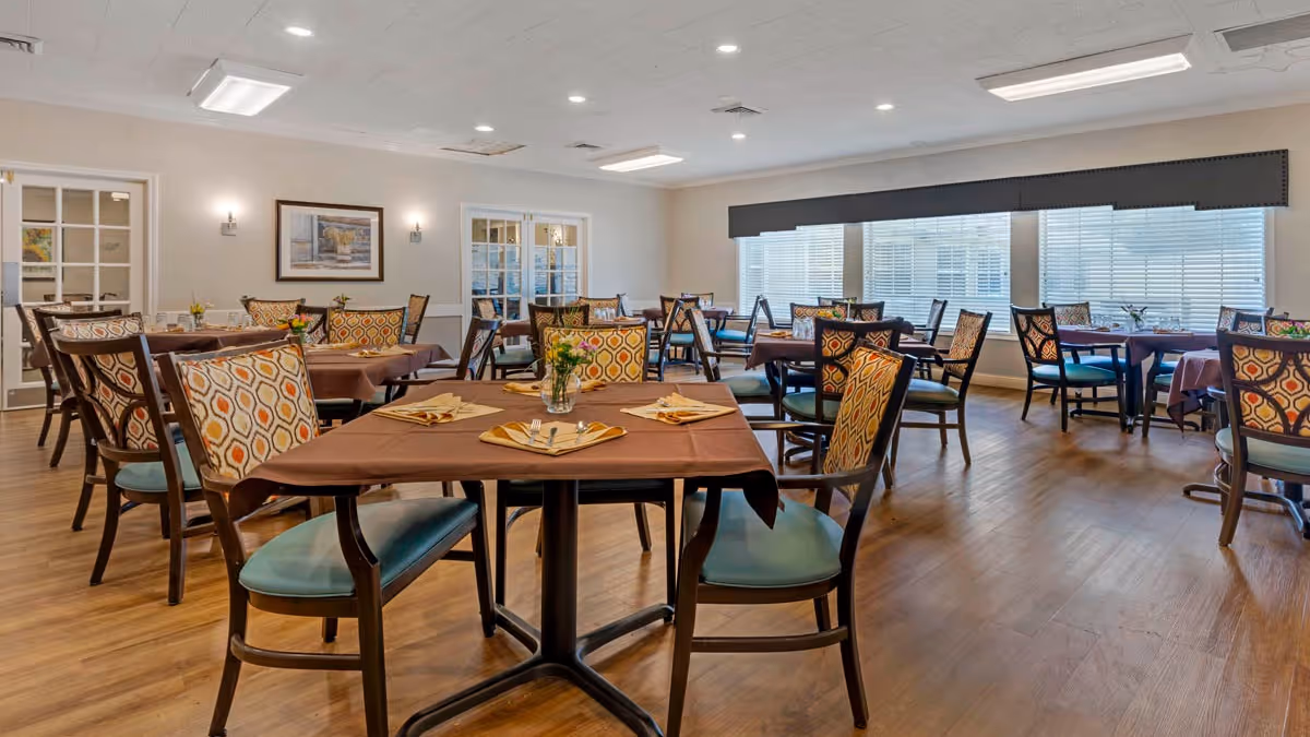 Spacious, well-lit dining room with multiple tables and patterned chairs set with napkins and small floral centerpieces.