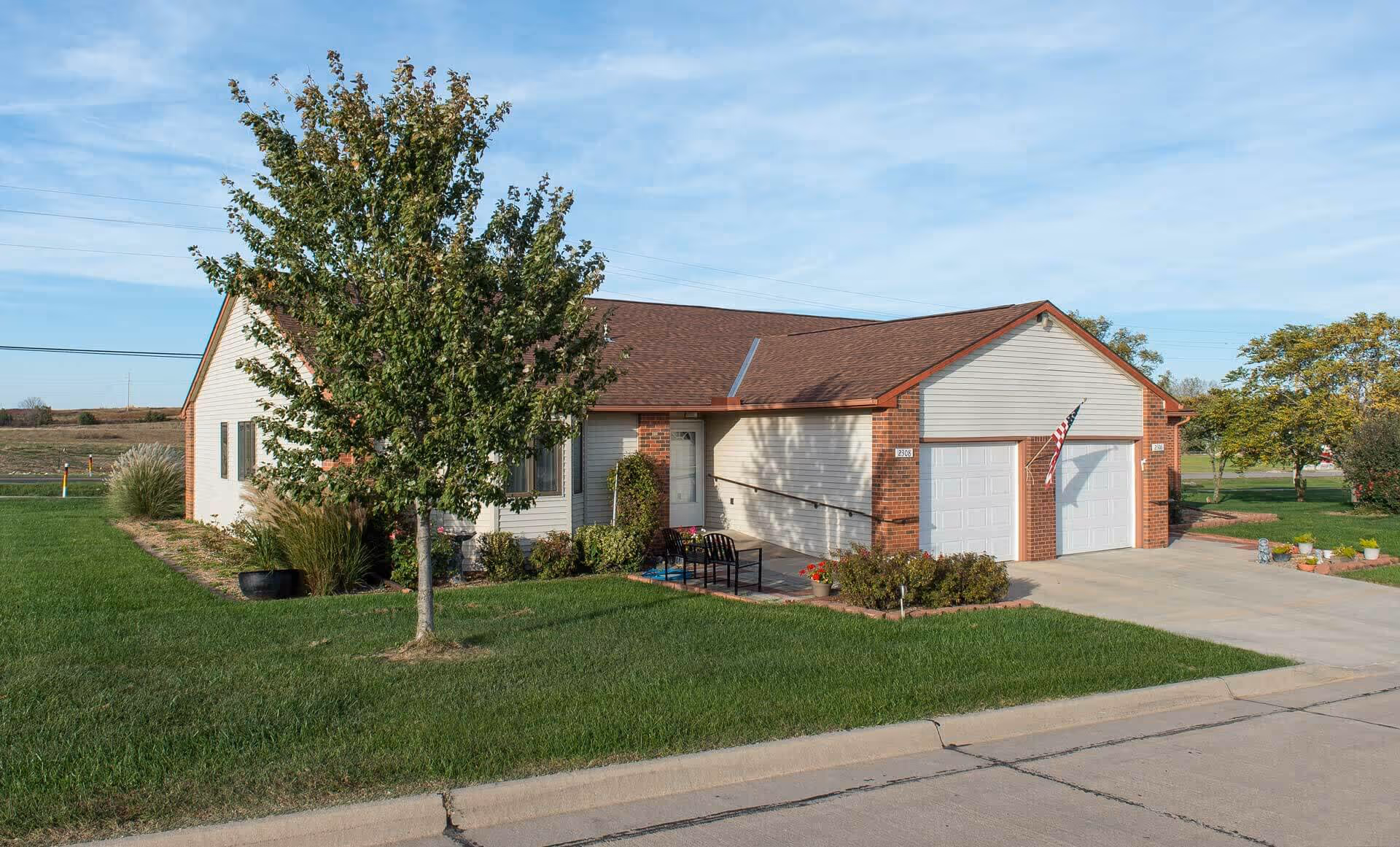 Single-story residential building with a brick and siding exterior, a two-car garage, an American flag mounted near the garage, a small patio with chairs, and a tree in the front yard under a partly cloudy sky.