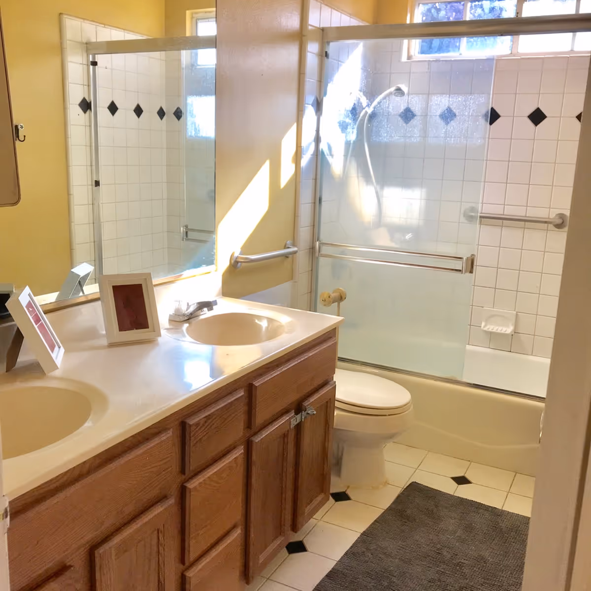 Well-lit bathroom with a double-sink vanity, toilet, and a tiled bathtub/shower with sliding glass doors.