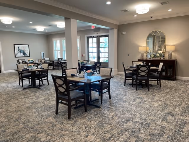 A spacious dining room with multiple dark wood tables and chairs arranged neatly on a patterned carpet. The room features soft lighting from ceiling fixtures and table lamps on a sideboard with a decorative mirror above it. Large windows and glass doors allow natural light to enter the room.