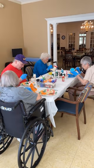Four elderly individuals seated around a table engaged in a craft activity involving painting. The table is covered with a white cloth and various art supplies including paint bottles and brushes are spread out. The setting appears to be a communal room with beige walls and a view into a dining area with multiple tables and chairs.
