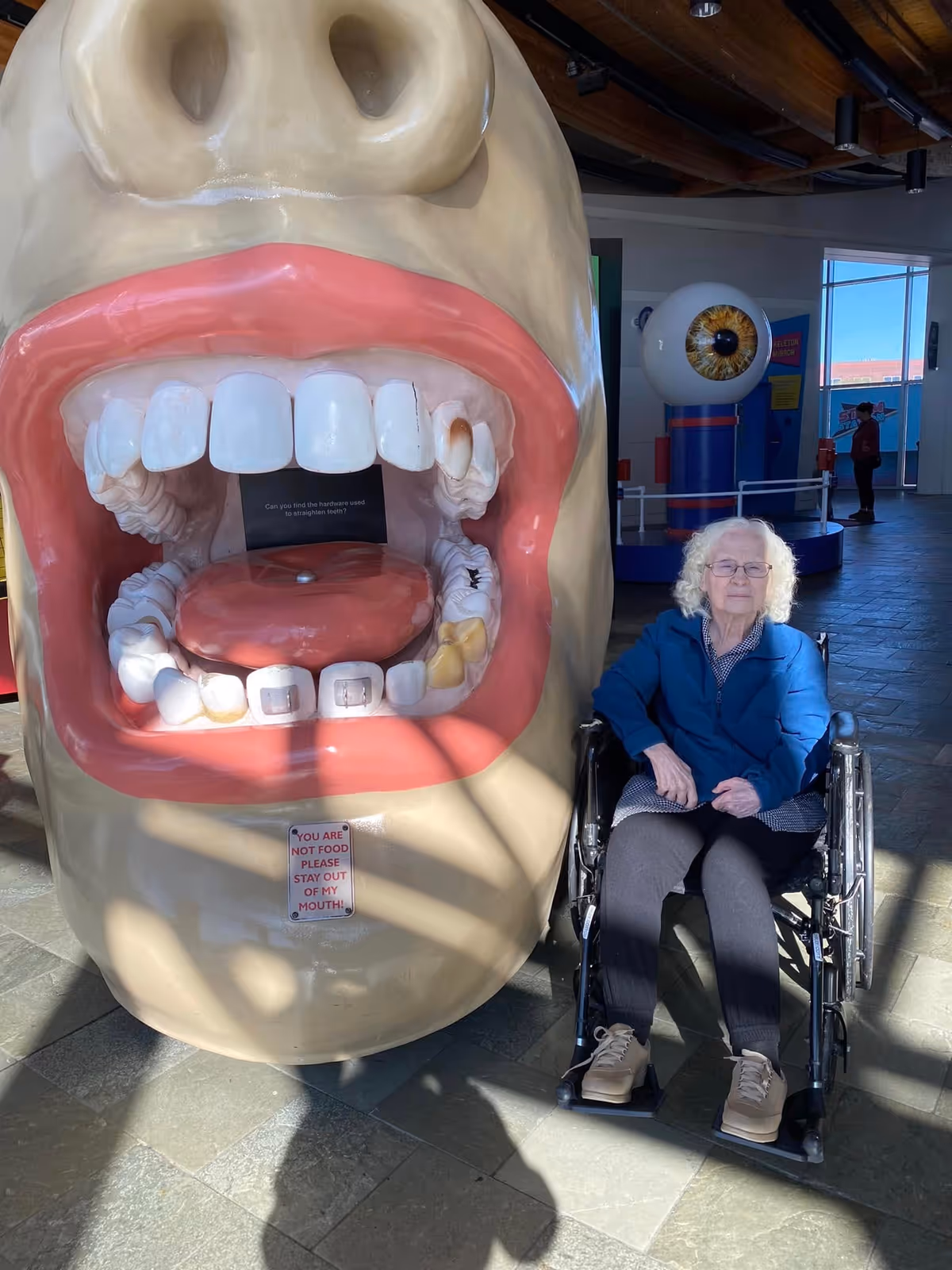 An elderly woman in a wheelchair sitting next to a large, open mouth model showing teeth and gums inside a public indoor space with a high ceiling and large windows.