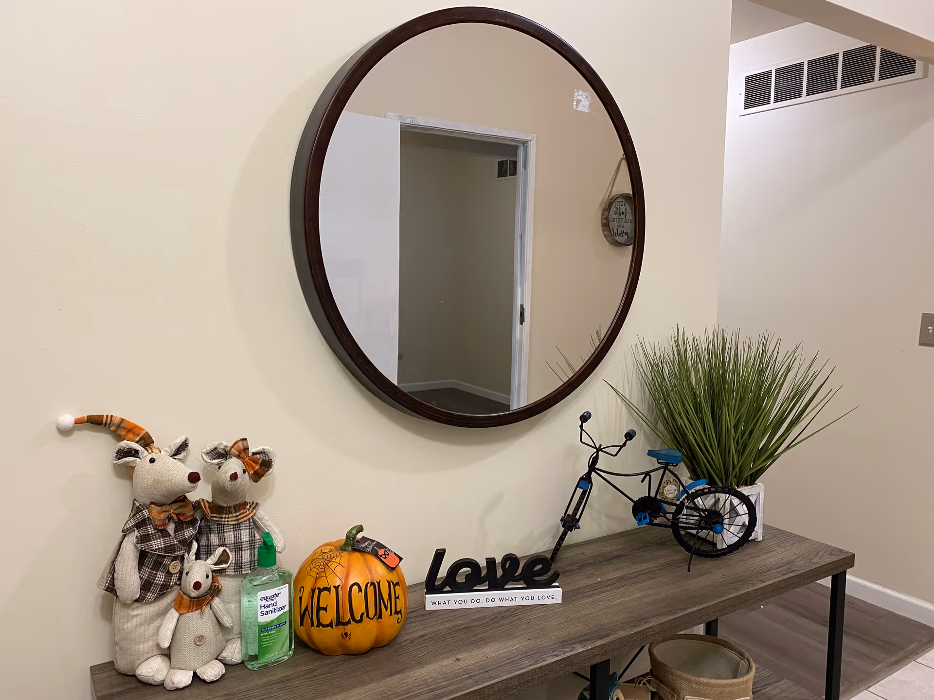 A wooden console table against a beige wall with a round mirror hanging above it. On the table are three stuffed animal decorations dressed in plaid, a bottle of hand sanitizer, a decorative pumpkin with the word 'WELCOME' written on it, a black metal bicycle sculpture, a green potted plant, and a decorative sign that says 'love' with the phrase 'WHAT YOU DO, DO WHAT YOU LOVE.' beneath it. The floor is tiled and there is a doorway reflected in the mirror.