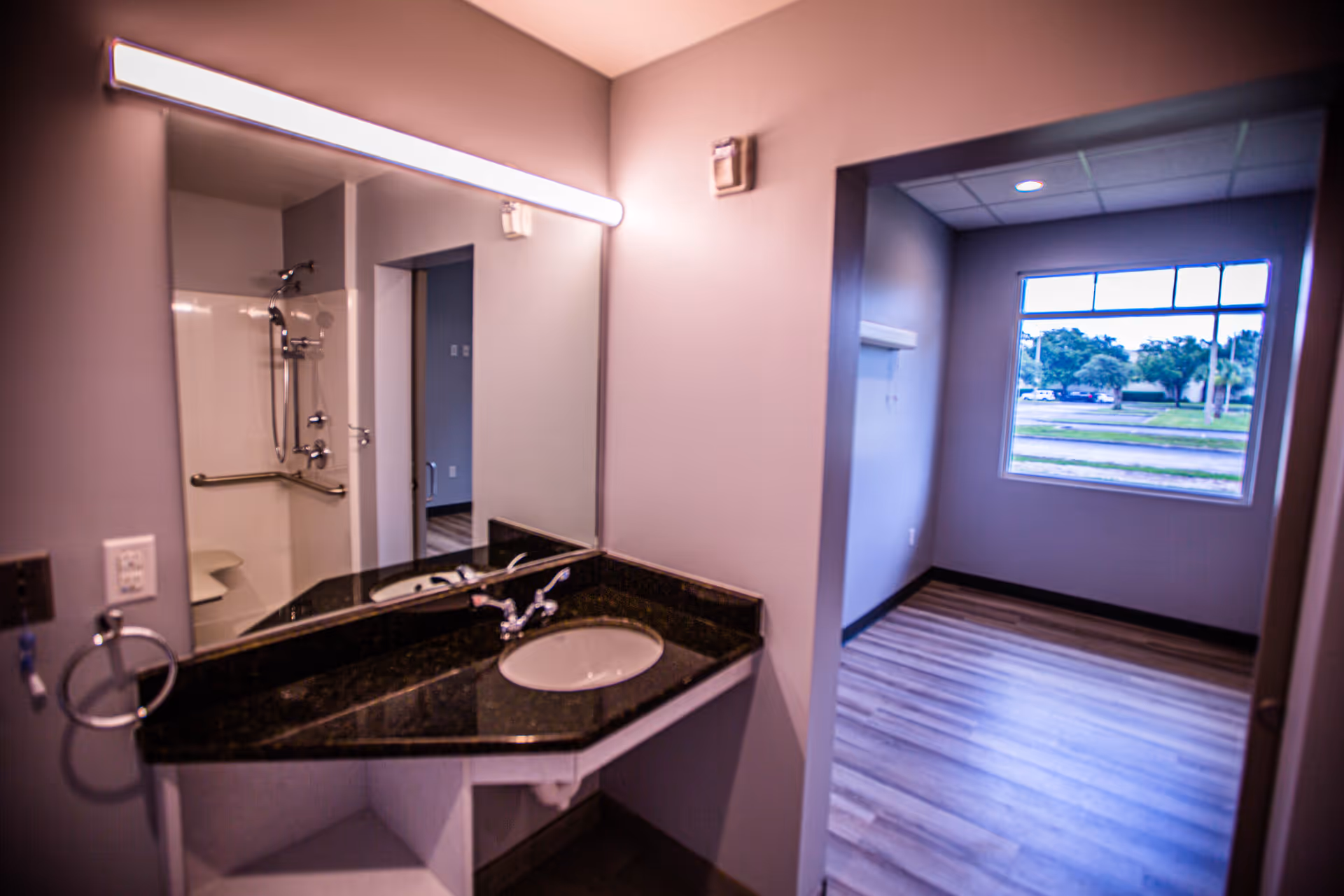 Interior view of a senior living facility bathroom area with a black granite countertop and sink, a large mirror with a horizontal light above it, and a walk-in shower with grab bars. Adjacent to the bathroom is an empty room with wood flooring and a large window showing an outdoor view with trees and parked cars.