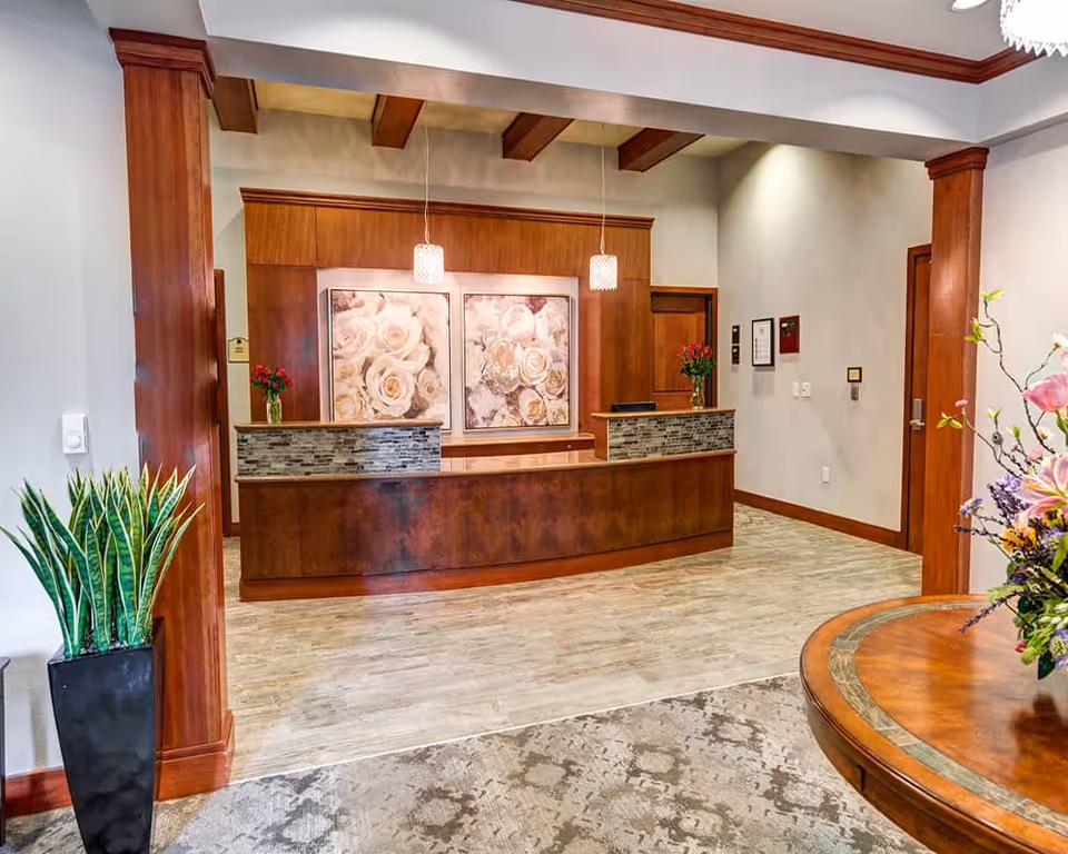 Reception area with a wooden front desk featuring stone accents, two pendant lights hanging from the ceiling, and floral artwork on the wall behind the desk. There are plants on either side of the desk and a round wooden table with a flower arrangement in the foreground.