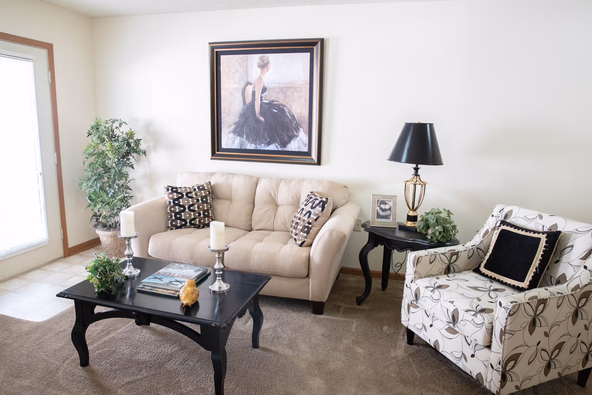 A cozy living room with a beige tufted sofa adorned with patterned pillows, a black wooden coffee table with candles, books, and a small decorative item, a patterned armchair with a black cushion, a black side table with a lamp, a framed photo, and a green plant. A large framed painting of a woman in a black dress hangs on the wall above the sofa. There is a potted plant near a glass door letting in natural light.