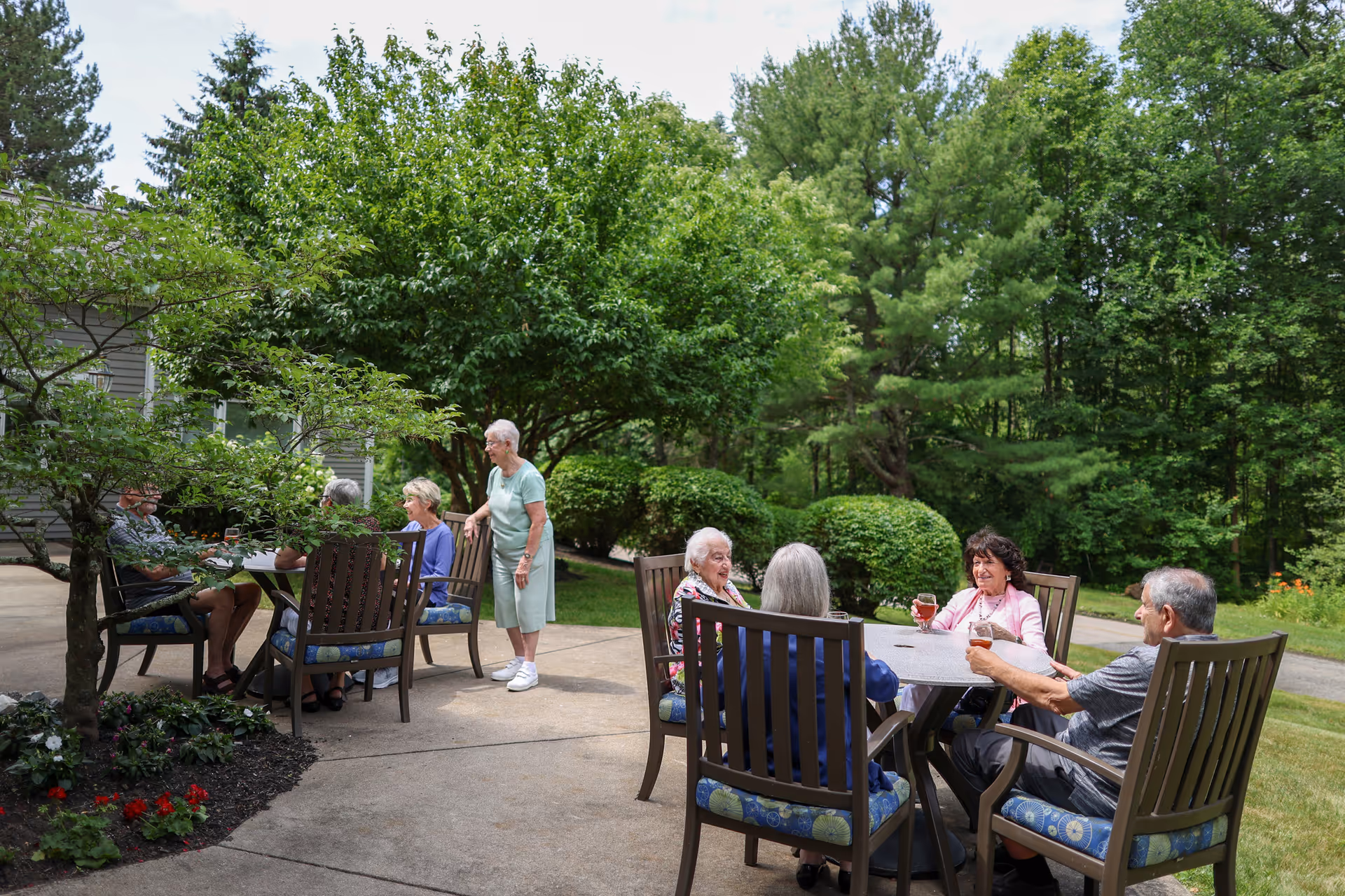 A group of elderly people sitting and standing around two outdoor tables on a patio surrounded by lush green trees and bushes, enjoying drinks and conversation on a sunny day.