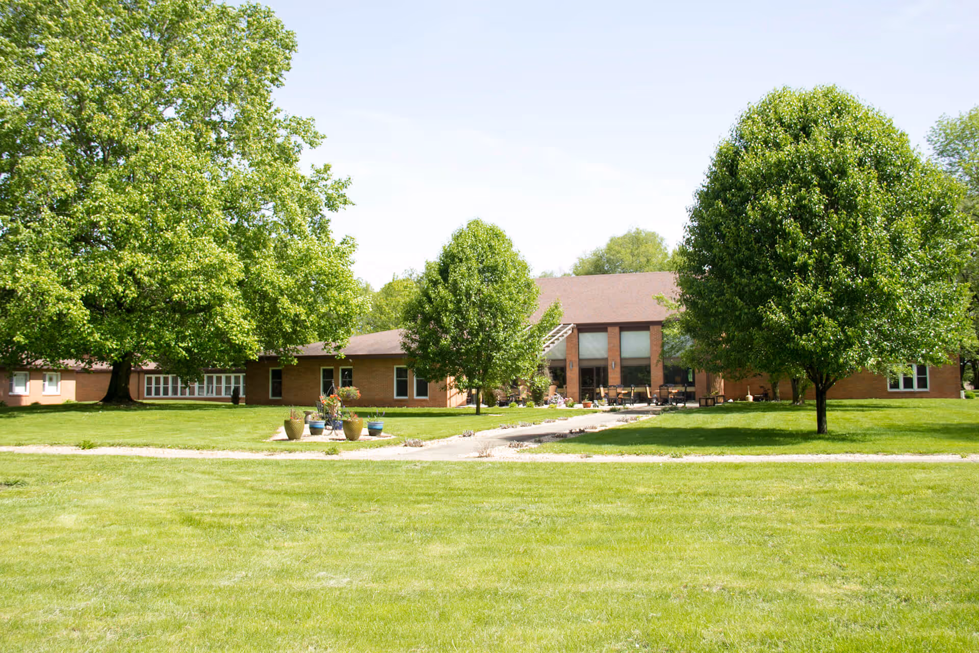 Exterior view of Friendship Hill Retirement Center showing a single-story brick building surrounded by green grass and several large leafy trees under a clear sky.
