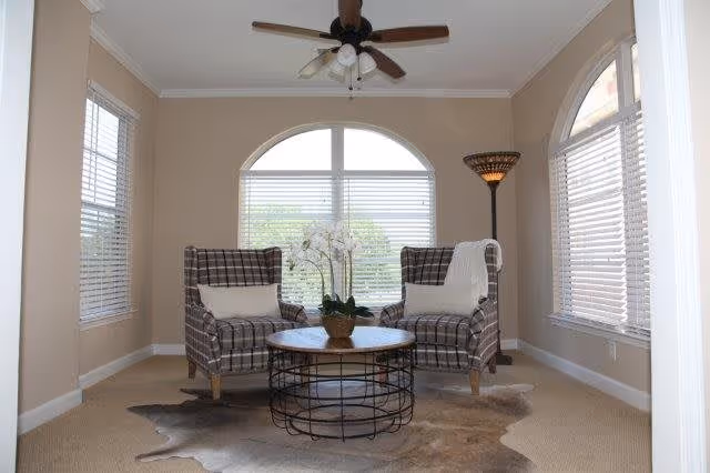 A cozy sitting area with two plaid armchairs facing each other, each with a white pillow and one with a white throw blanket. Between the chairs is a round coffee table with a wireframe base and a wooden top, holding a potted orchid plant. The room has beige walls, carpeted floor with a cowhide rug, three large windows with white blinds, a ceiling fan with lights, and a tall floor lamp in the corner.
