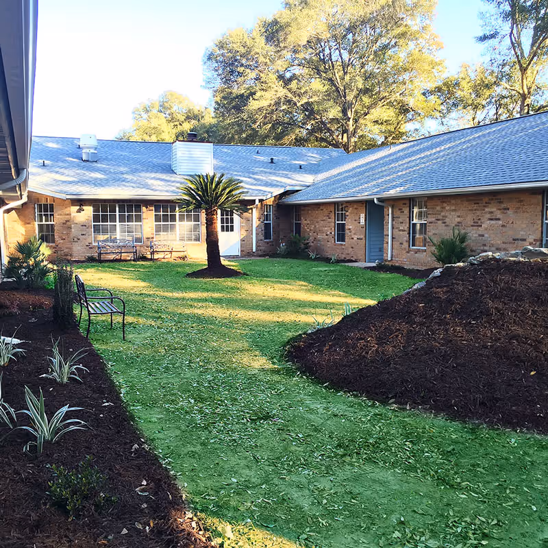 A sunny courtyard area with green grass, a small palm tree in the center, and a few benches along the edges. The courtyard is surrounded by a single-story brick building with several windows and doors. There are landscaped flower beds with mulch and plants along the sides.