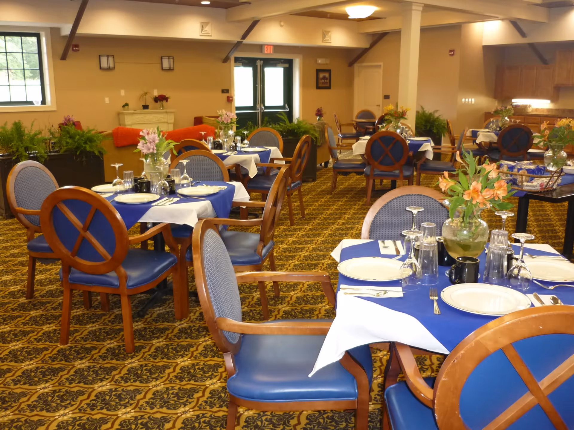A dining room in a senior living facility with tables covered in blue and white tablecloths, set with plates, glasses, silverware, and floral centerpieces. The room has patterned carpet, wooden chairs with blue cushions, and plants along the walls. There is a double door exit at the back and warm lighting overhead.