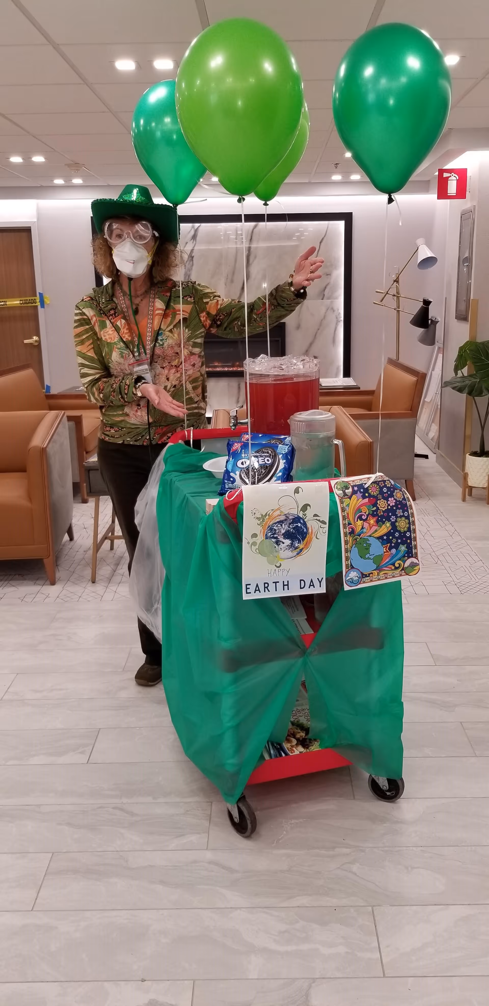 A person wearing a green hat, protective goggles, and a face mask stands behind a decorated cart with green balloons attached. The cart is covered with a green cloth and has Earth Day themed decorations, including a sign that says 'Happy Earth Day'. The setting appears to be a common area with brown chairs and a fireplace in the background.