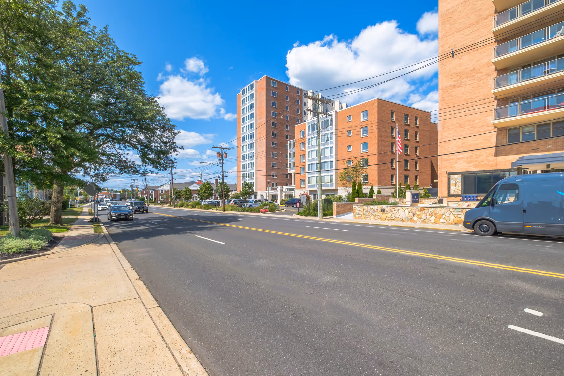 Wide street view showing a multi-story brick residential building front with parked vehicles, sidewalk, and trees under a blue sky.