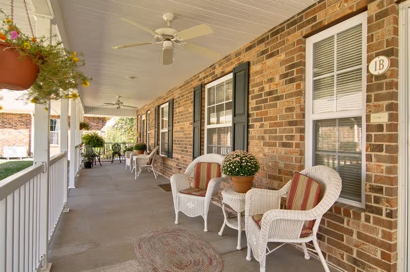 A covered outdoor porch area with white wicker chairs featuring red and beige striped cushions, small tables with potted plants, ceiling fans, and hanging flower pots. The porch is attached to a brick building with windows and black shutters.