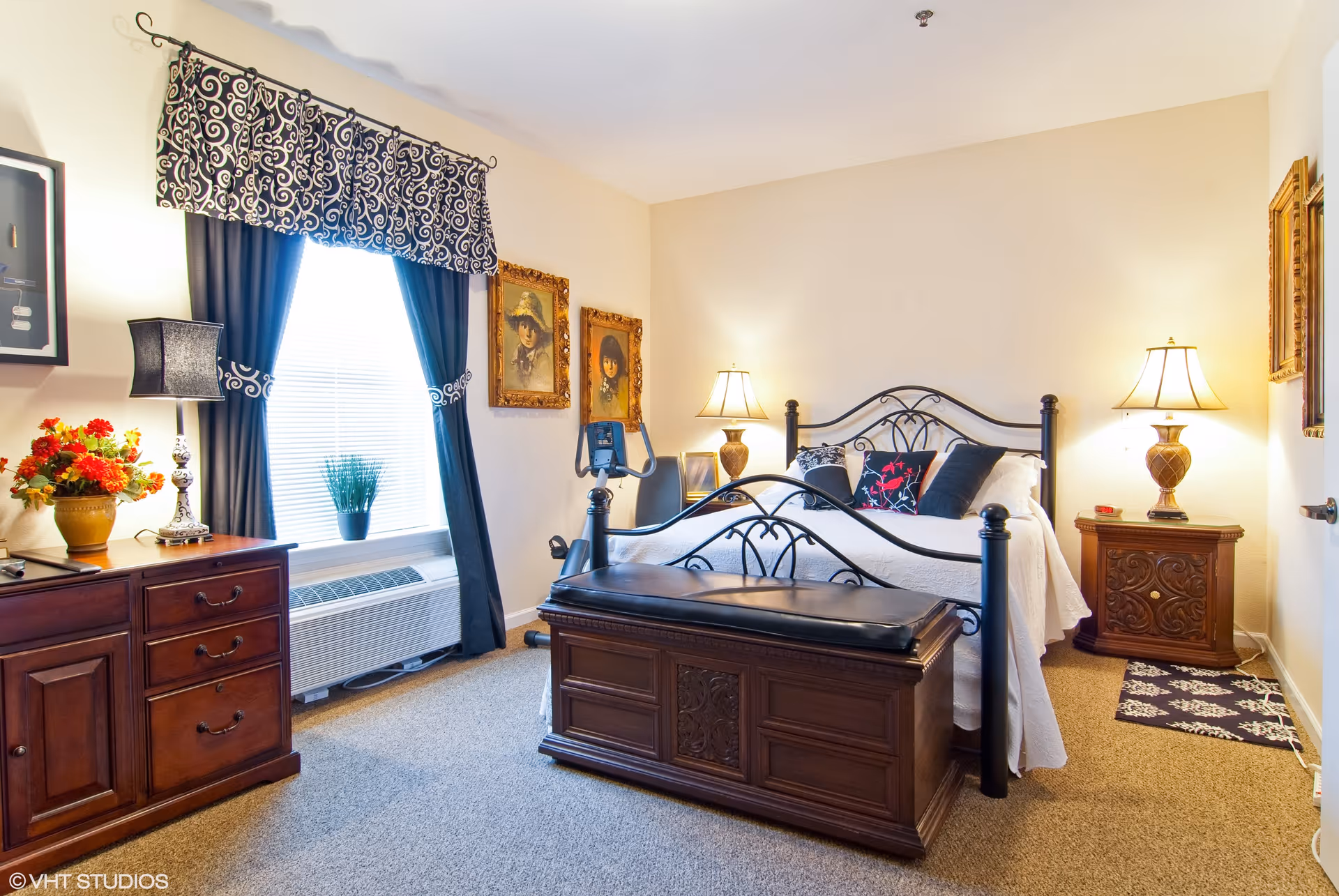A well-lit bedroom featuring a large bed with a decorative black metal frame and white bedding. There are two bedside tables with matching lamps on either side of the bed. A wooden chest with a cushioned top is placed at the foot of the bed. The room has beige walls, carpeted floor, and a window with dark curtains and a patterned valance. Two framed portraits hang on the wall beside the bed, and a small exercise bike is positioned near the window.