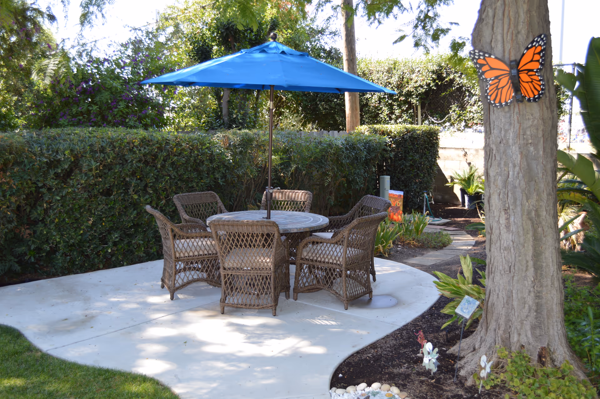 Outdoor patio area with a round table and six wicker chairs under a large blue umbrella, surrounded by greenery and a tree with a decorative orange butterfly attached to its trunk.