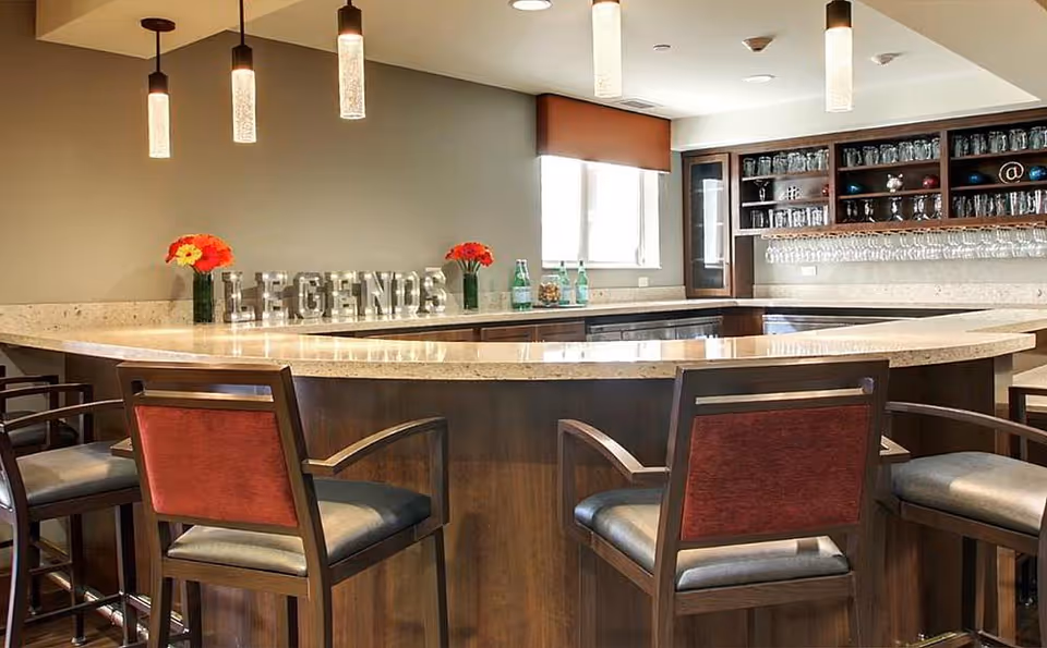 Interior view of a senior living facility bar area with a curved marble countertop, wooden bar stools with red and gray cushions, hanging pendant lights, shelves with glassware, and decorative items including flowers and a sign that reads 'LEGENDS'.