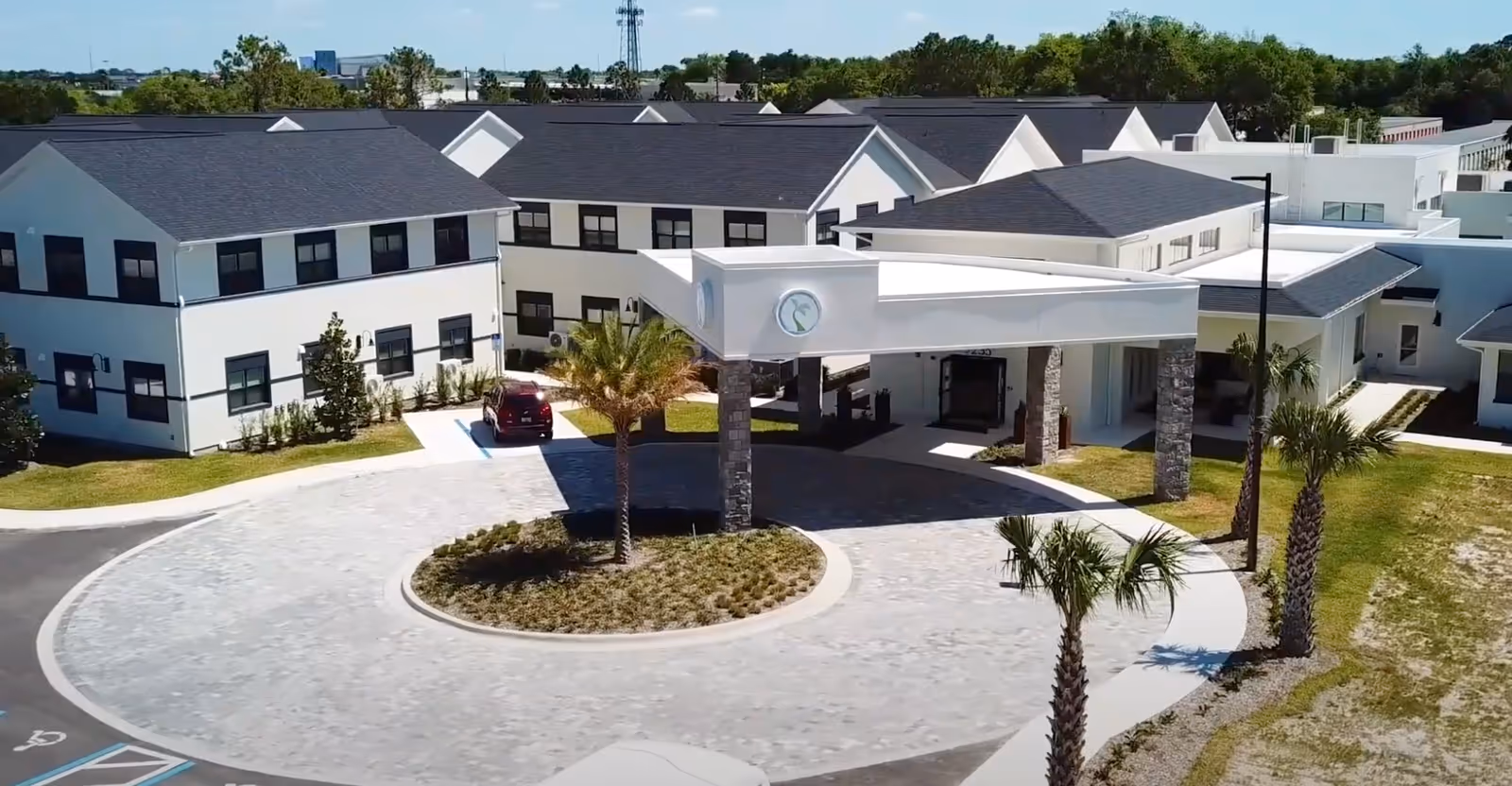 Exterior view of Strive at Fern Park senior living community showing a circular driveway with palm trees and a covered entrance supported by stone pillars. The building has white walls and dark roofs with multiple windows.