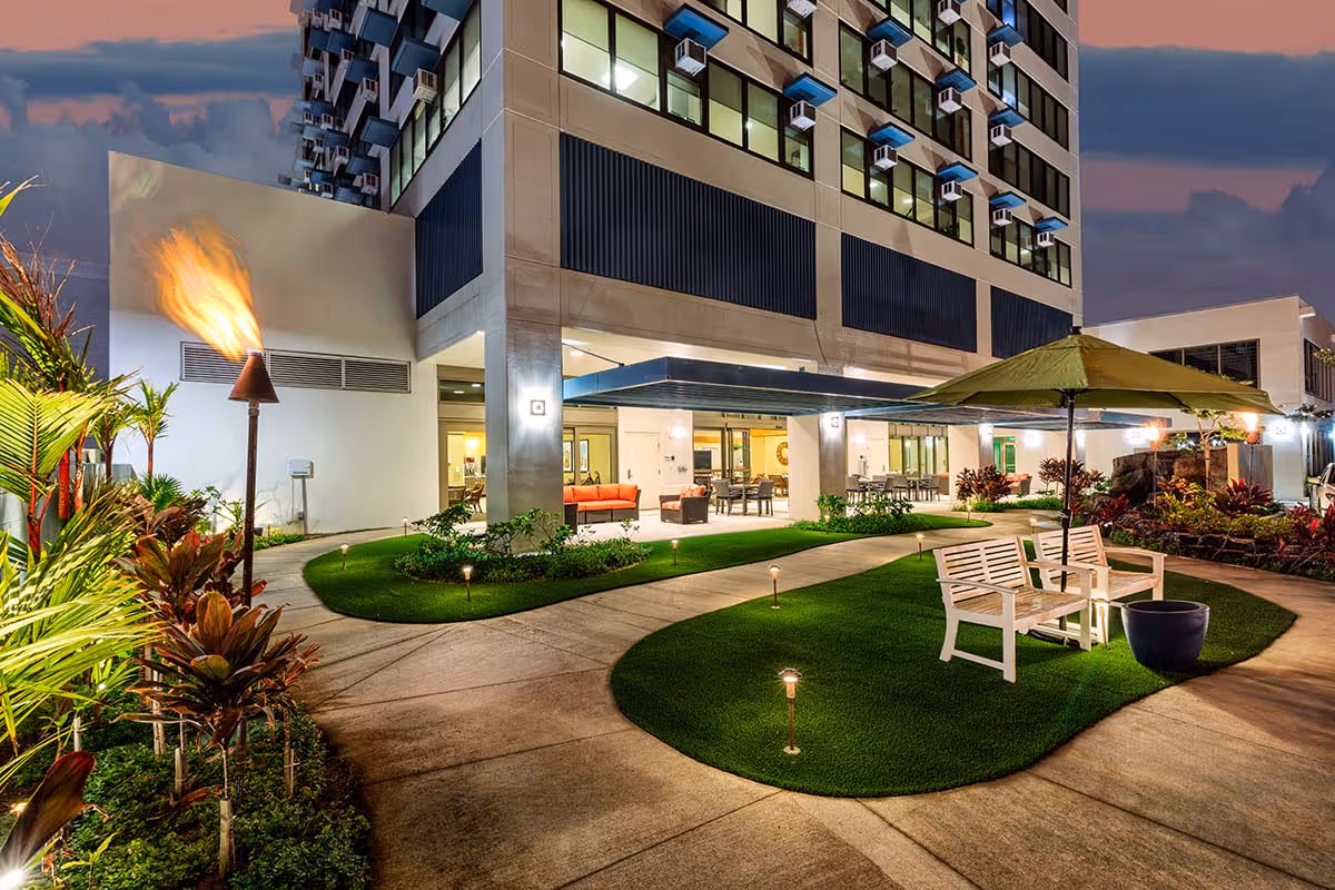 Outdoor seating area at night with benches, a large umbrella, tiki torch, and landscaped greenery in front of a multi-story building with illuminated windows.