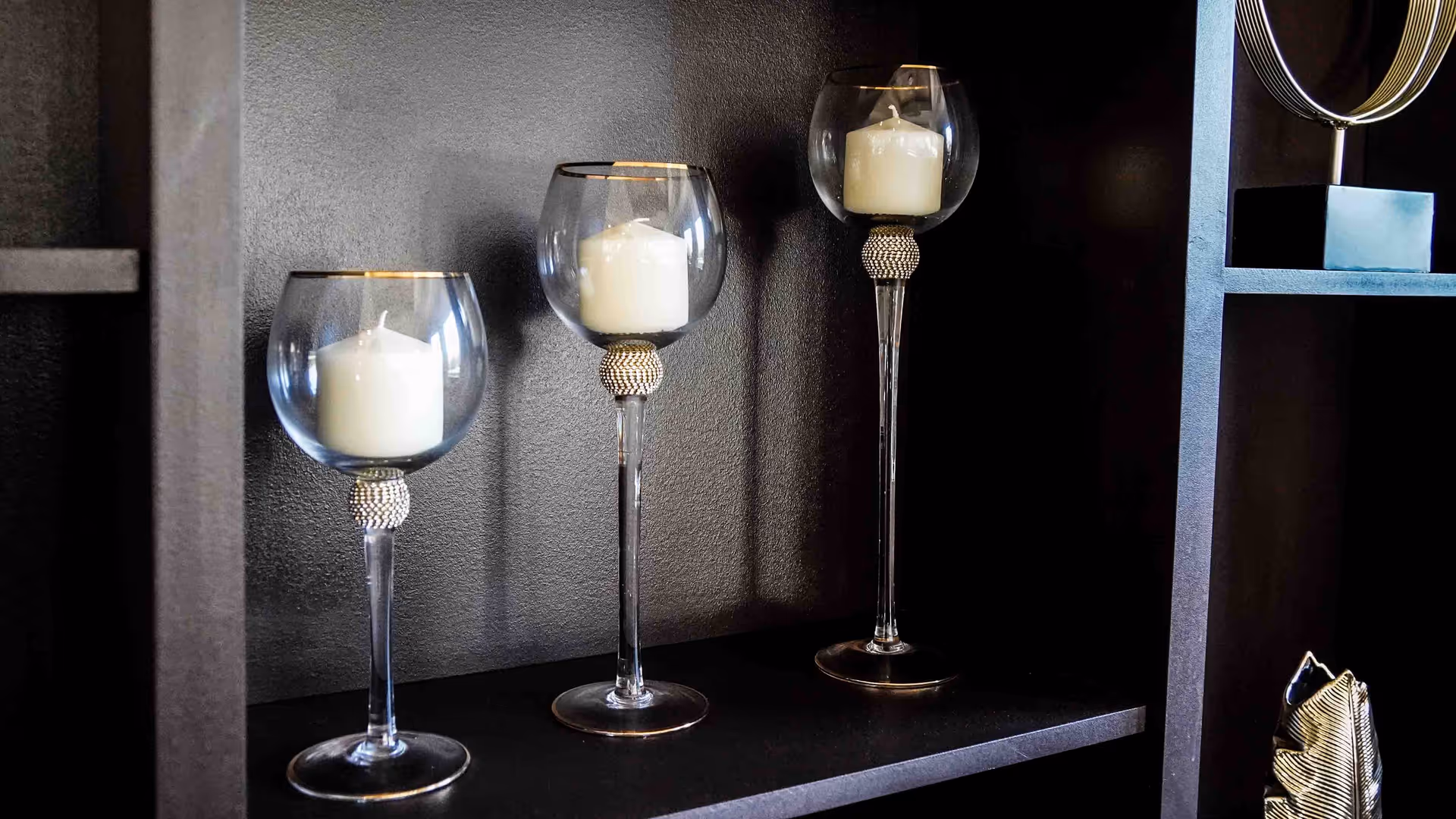Three decorative glass candle holders of varying heights, each containing a white candle, displayed on a dark shelf against a dark wall.