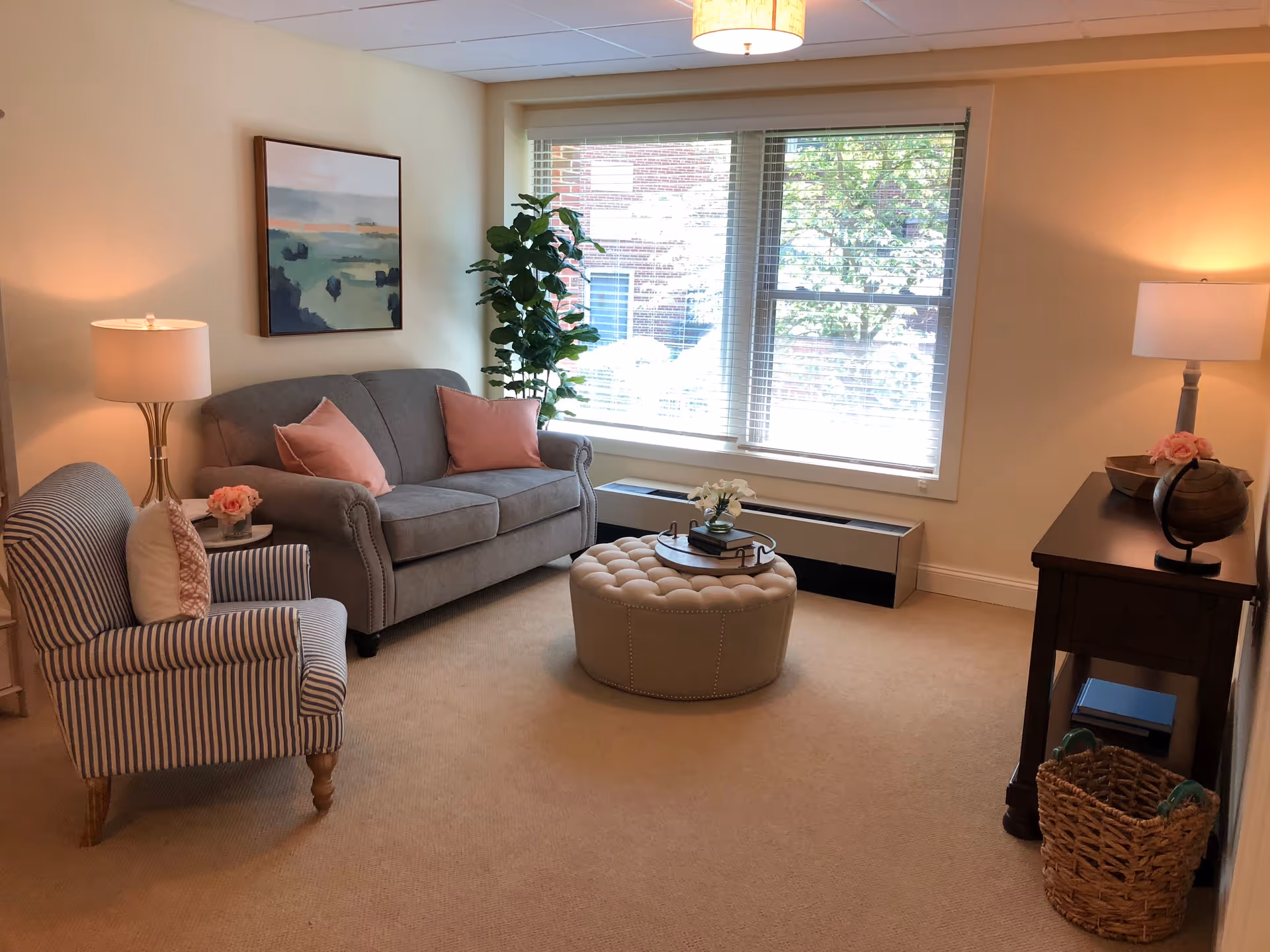 A cozy living room with a gray sofa adorned with pink pillows, a striped armchair with a white pillow, a round tufted ottoman in the center, and a wooden side table with a lamp and decorative items. Large windows with blinds allow natural light to fill the room, and a potted plant and framed artwork add to the decor.