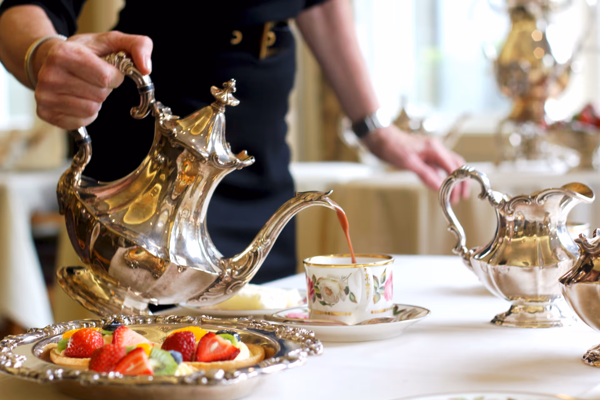 A person pouring tea from an ornate silver teapot into a floral teacup on a white tablecloth. In the foreground, there is a silver tray with fruit tarts topped with strawberries, kiwi, and other fruits. Additional silver serving pieces are visible on the table.