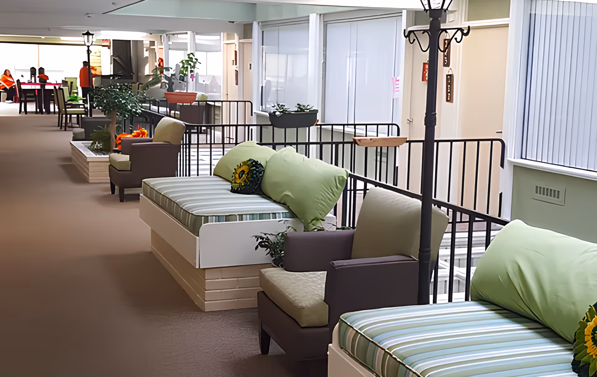 Indoor hallway seating area with cushioned benches and armchairs, decorated with green pillows and plants, featuring a railing and lamp posts along the corridor.