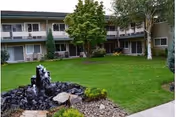 Grassy courtyard with a small rock fountain surrounded by a two-story residential building and trees.