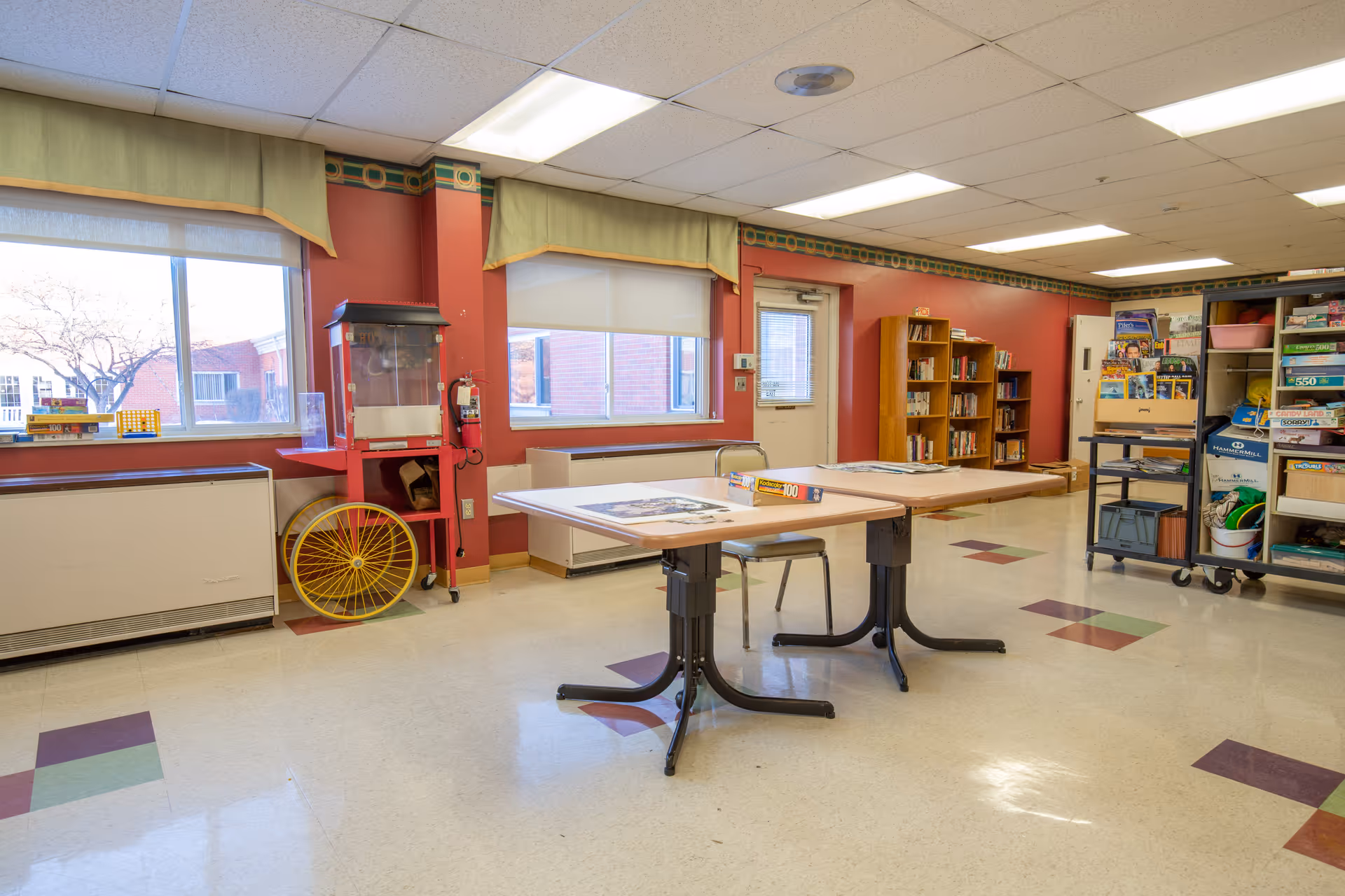 A spacious activity room with red walls and large windows letting in natural light. The room contains two tables with chairs, a popcorn machine, bookshelves filled with books, and a cart with various games and supplies. The floor has a pattern of colored tiles, and there is a door leading outside.