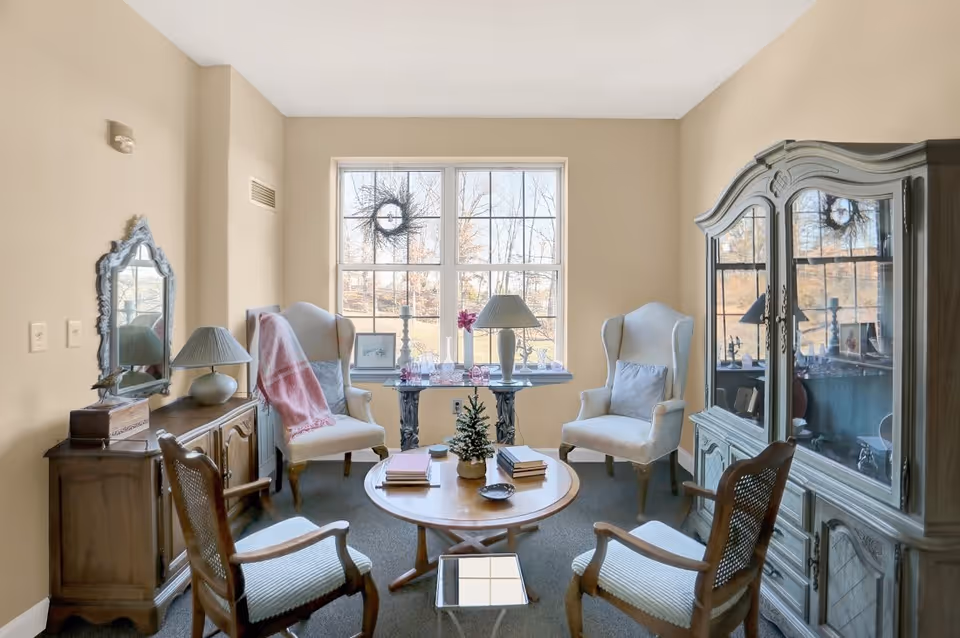 A cozy living room with four chairs arranged around a round wooden coffee table. The room features a large window with a view of trees outside, a wooden cabinet with a mirror and lamp on the left, and a large glass-fronted cabinet on the right. The walls are painted beige and the room is softly lit with natural light.