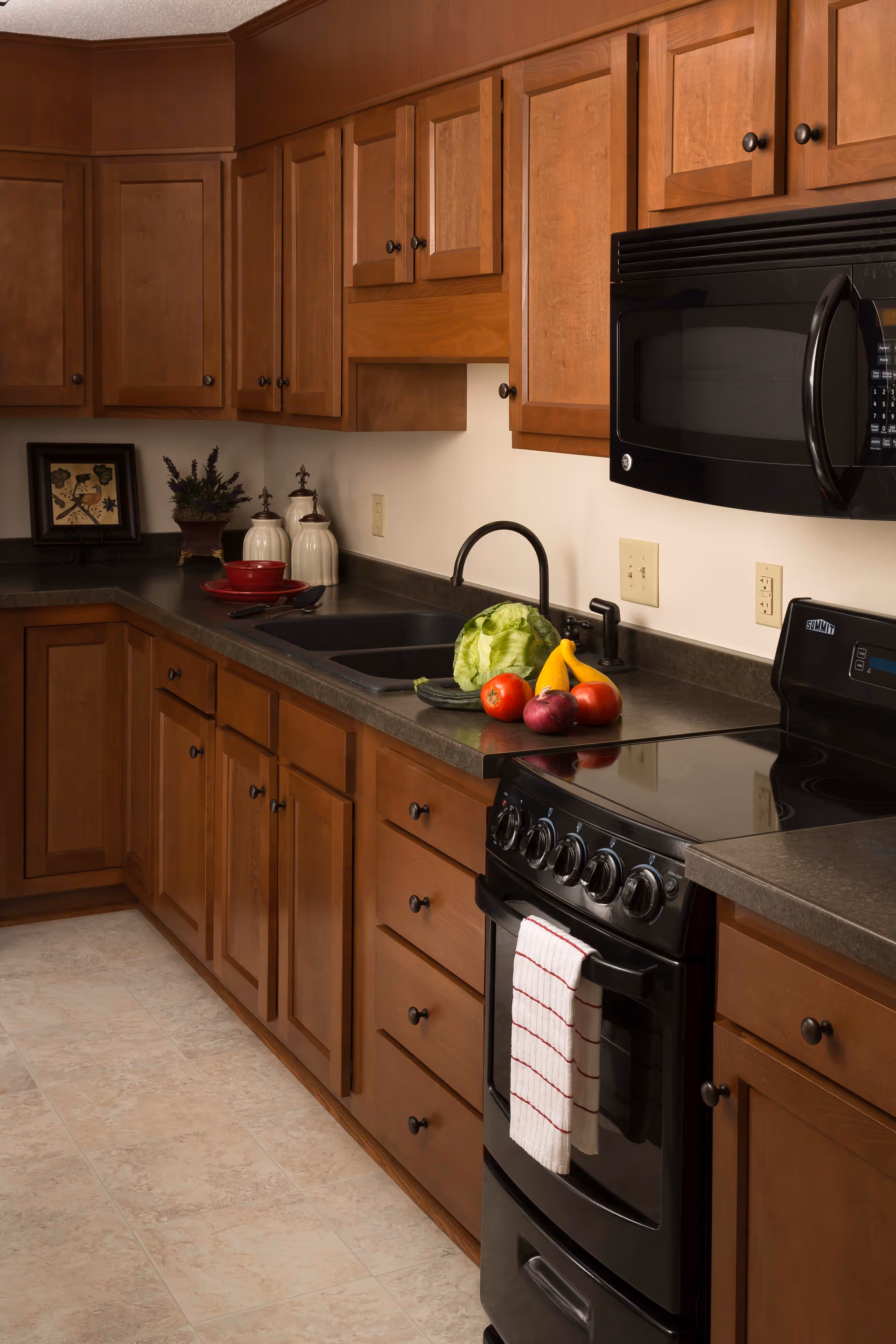 A kitchen with wooden cabinets, a black stove with a white and red striped towel hanging on the handle, a black microwave above the stove, a black sink with a faucet, and various vegetables including lettuce, tomatoes, a cucumber, a red onion, and yellow squash on the countertop. There are also decorative jars and a framed picture on the counter in the corner.