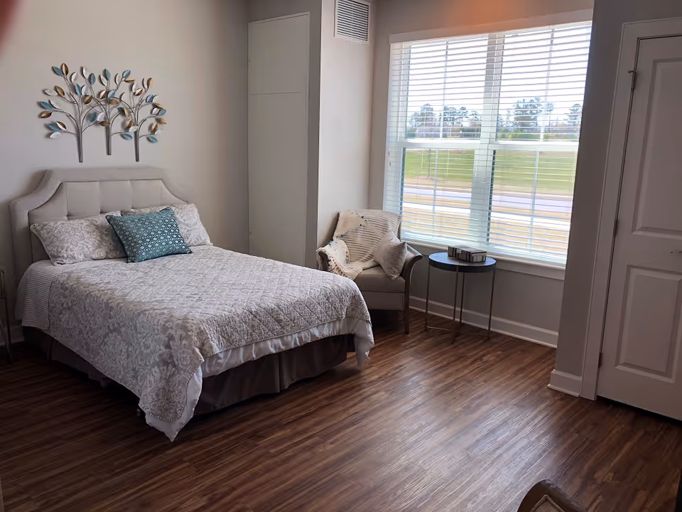 Well-lit bedroom with a bed, decorative metal wall art, an armchair and side table by a large window, and wood-look flooring.