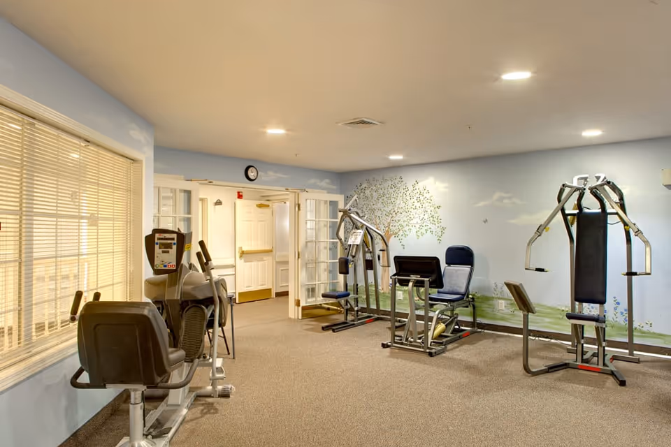 A small fitness room in a senior living facility with exercise machines including a recumbent bike, a leg press, and a multi-function weight machine. The walls are painted light blue with a mural of a tree and clouds. There is a window with blinds on the left and double doors leading to a hallway in the background.