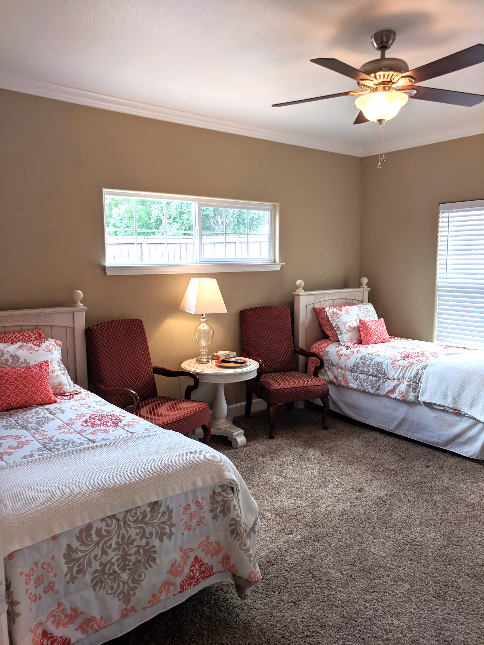 Two twin beds with patterned bedding, a small round table with a lamp and two chairs in a carpeted bedroom with a ceiling fan.