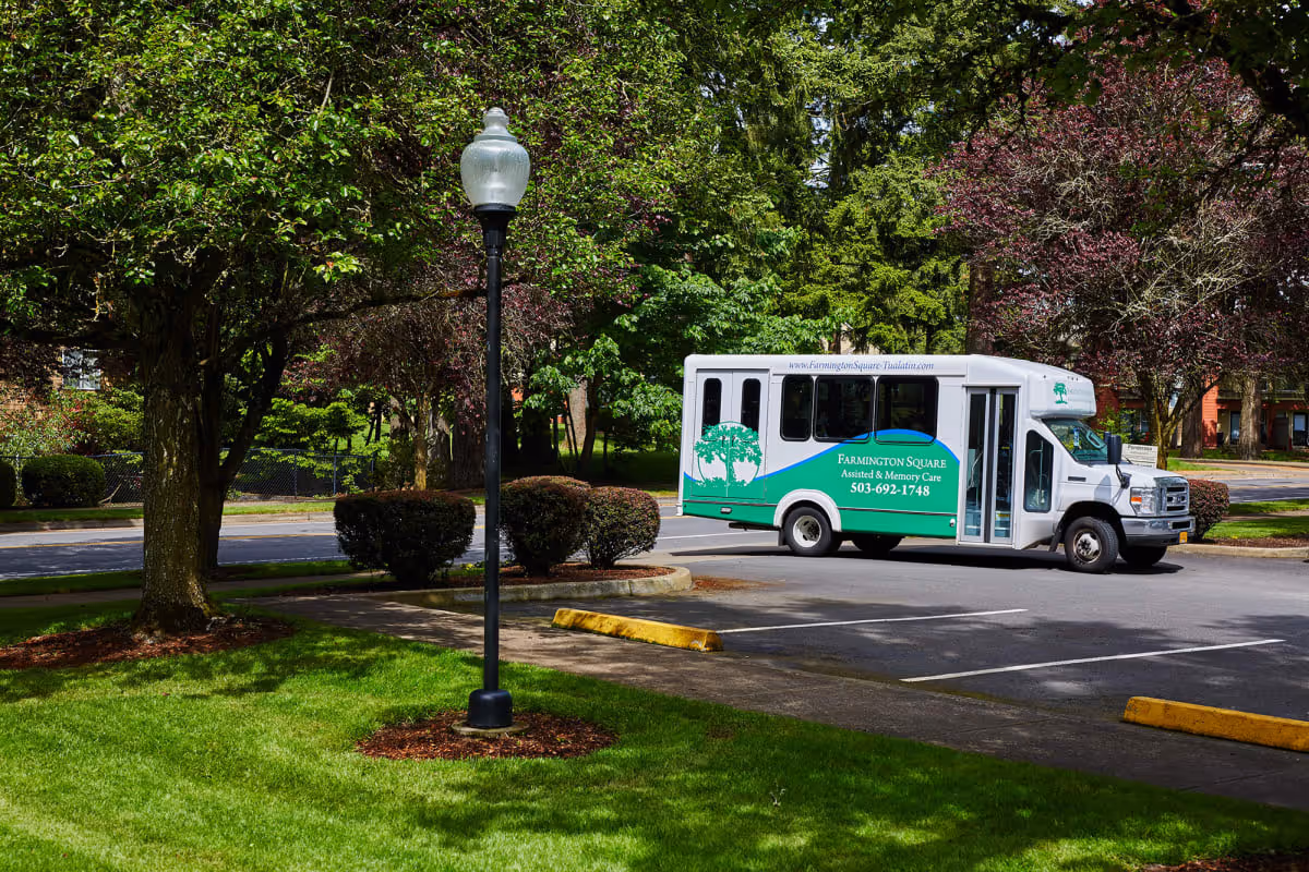A white and green senior living shuttle bus parked in a tree-lined parking lot next to a lamp post and manicured lawn.
