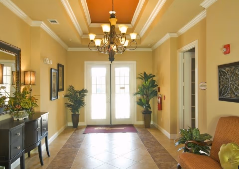 A warmly lit interior hallway with beige walls and tiled floor leading to double glass doors. The ceiling features a decorative orange inset with a chandelier. There are potted plants on either side of the doors, a dark wooden console table with a mirror and lamp on the left, and an upholstered chair with a green pillow on the right.